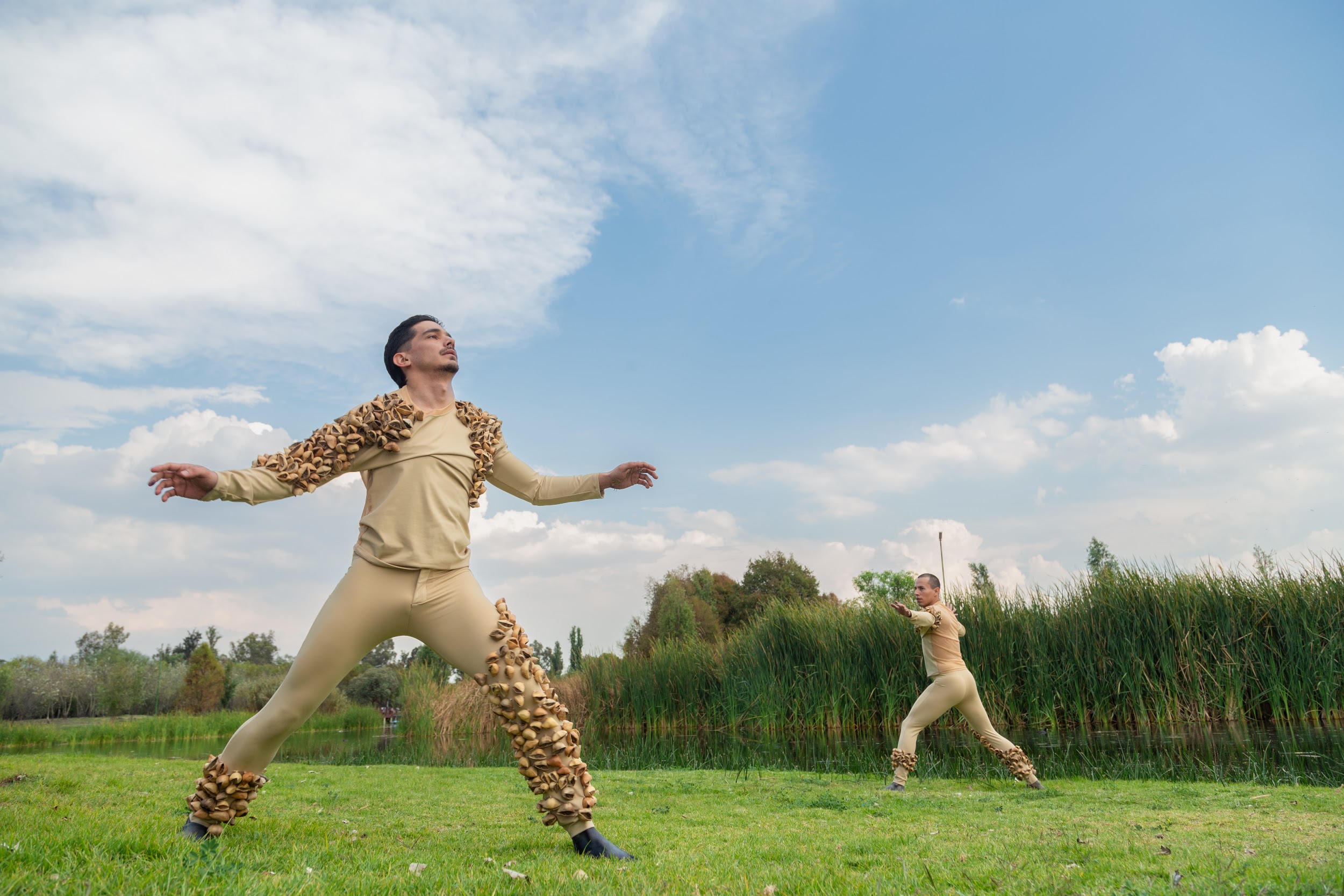 Two men in beige costumes with brown seed-like attachments perform expressive dance poses outdoors near a pond and tall grass.