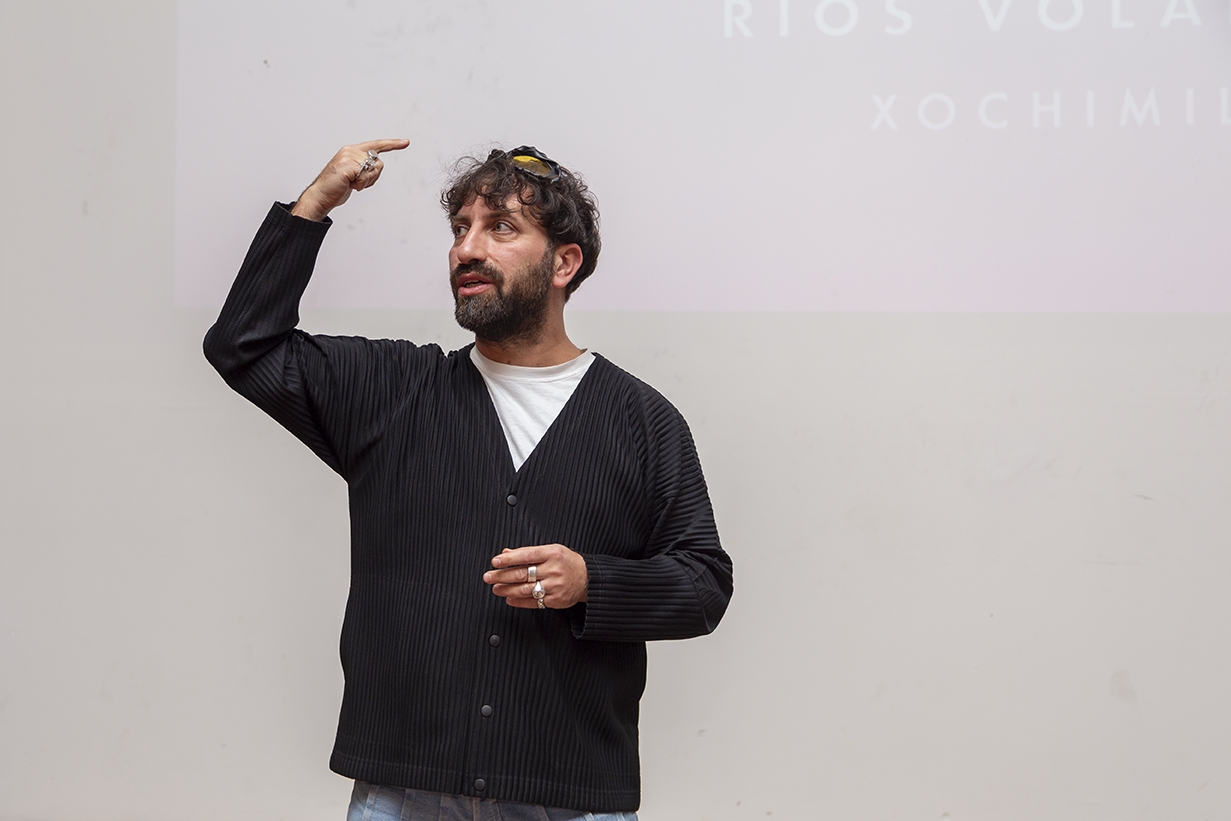 Man with curly hair and beard wearing a black jacket and white shirt, gesturing with his right hand near his head.