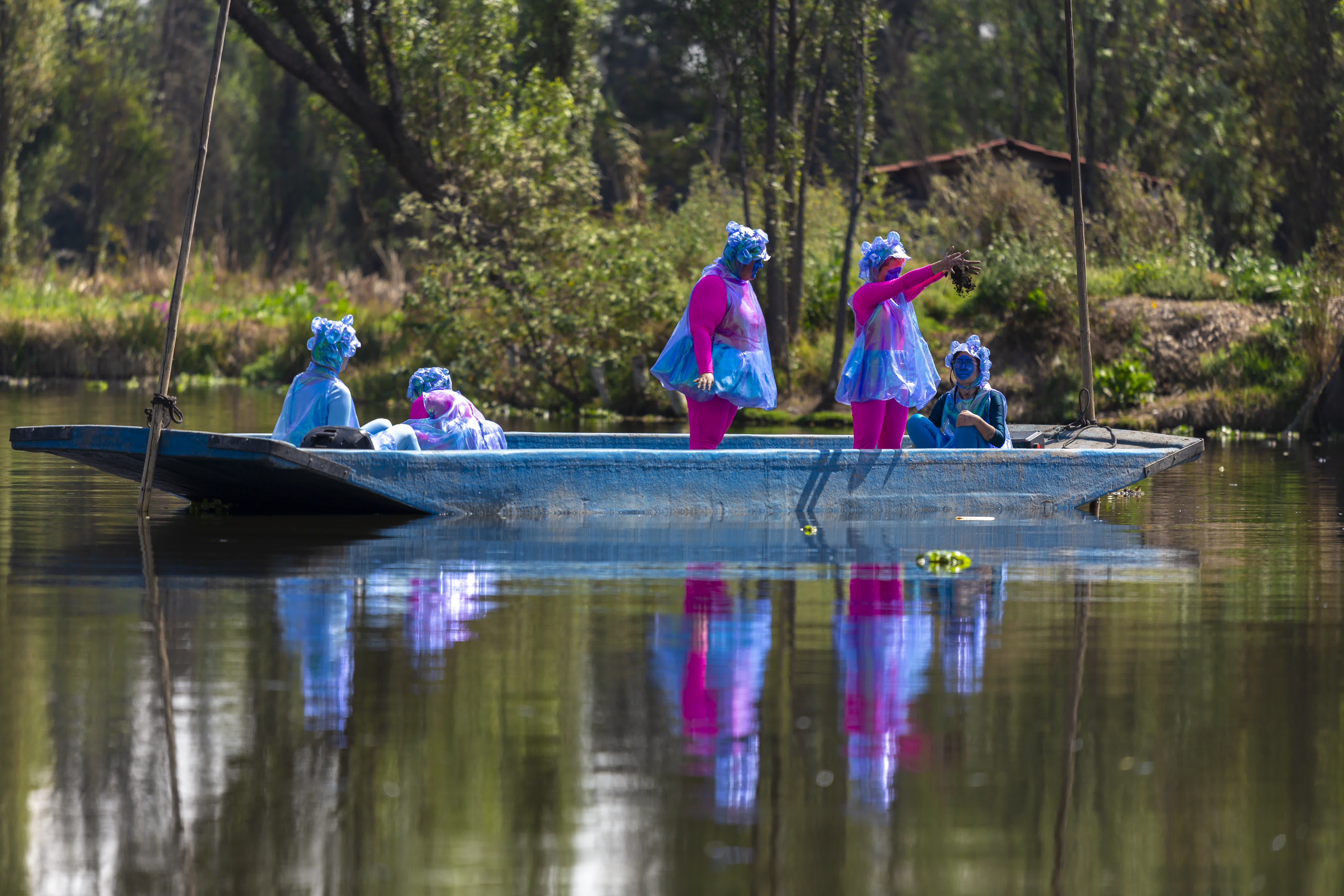 A group of seven people dressed in bright blue and pink costumes standing and sitting in a blue boat on calm water with trees in the background.
