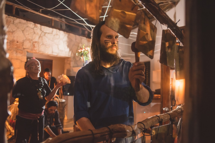 Bearded man with face paint playing metal percussion bars indoors with other musicians in the background.