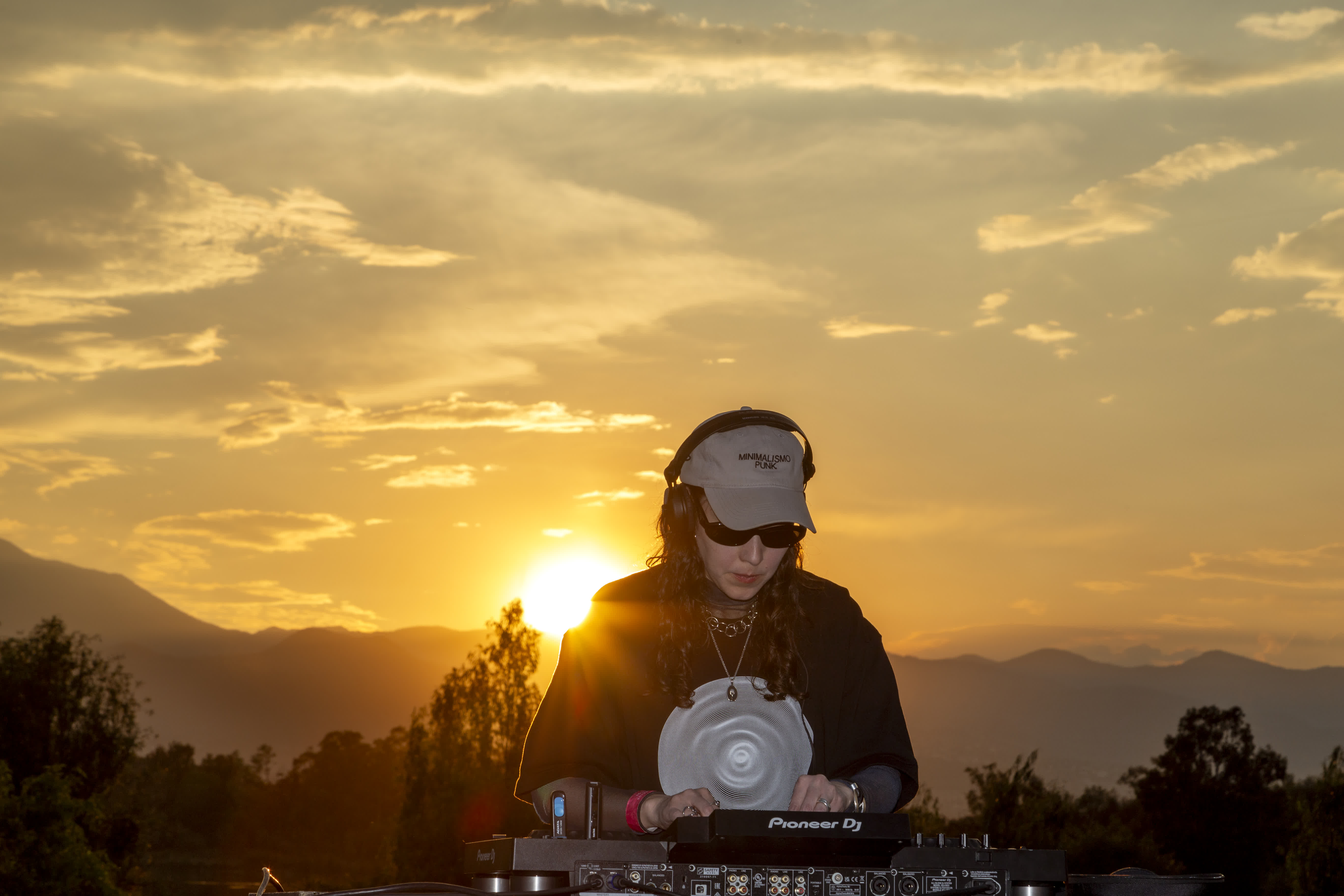 DJ wearing headphones and a cap playing on Pioneer DJ equipment outdoors at sunset.