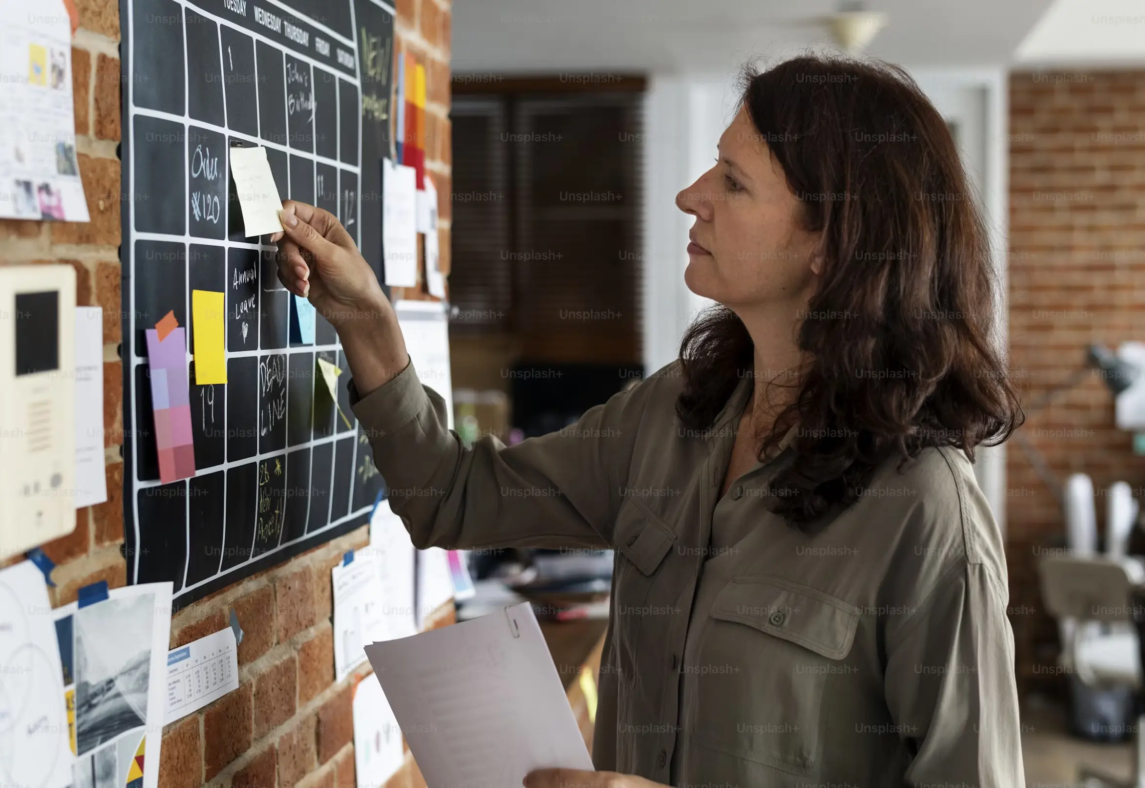 Woman placing a sticky note on a blackboard calendar pinned on a brick wall while holding papers.