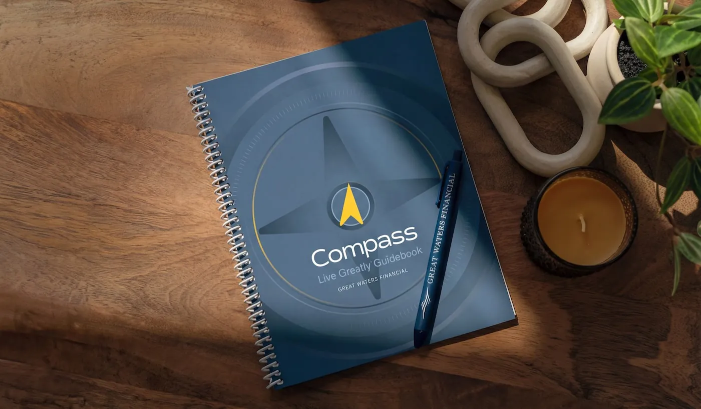 Spiral-bound book titled 'Create Your Compass Life Design Guidebook' placed on a wooden table next to a candle, decorative object, and a small potted plant.