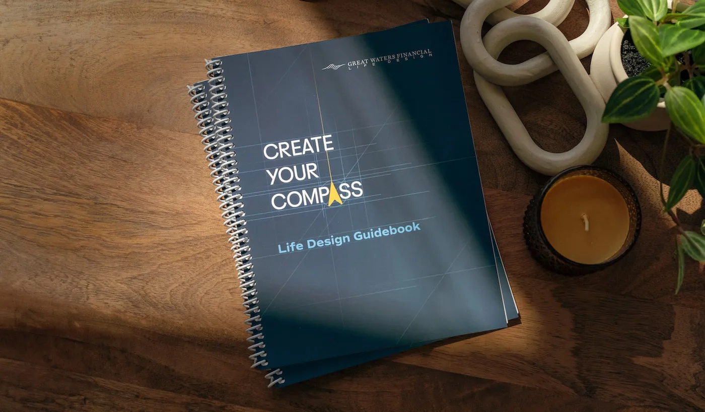 Spiral-bound book titled 'Create Your Compass Life Design Guidebook' placed on a wooden table next to a candle, decorative object, and a small potted plant.