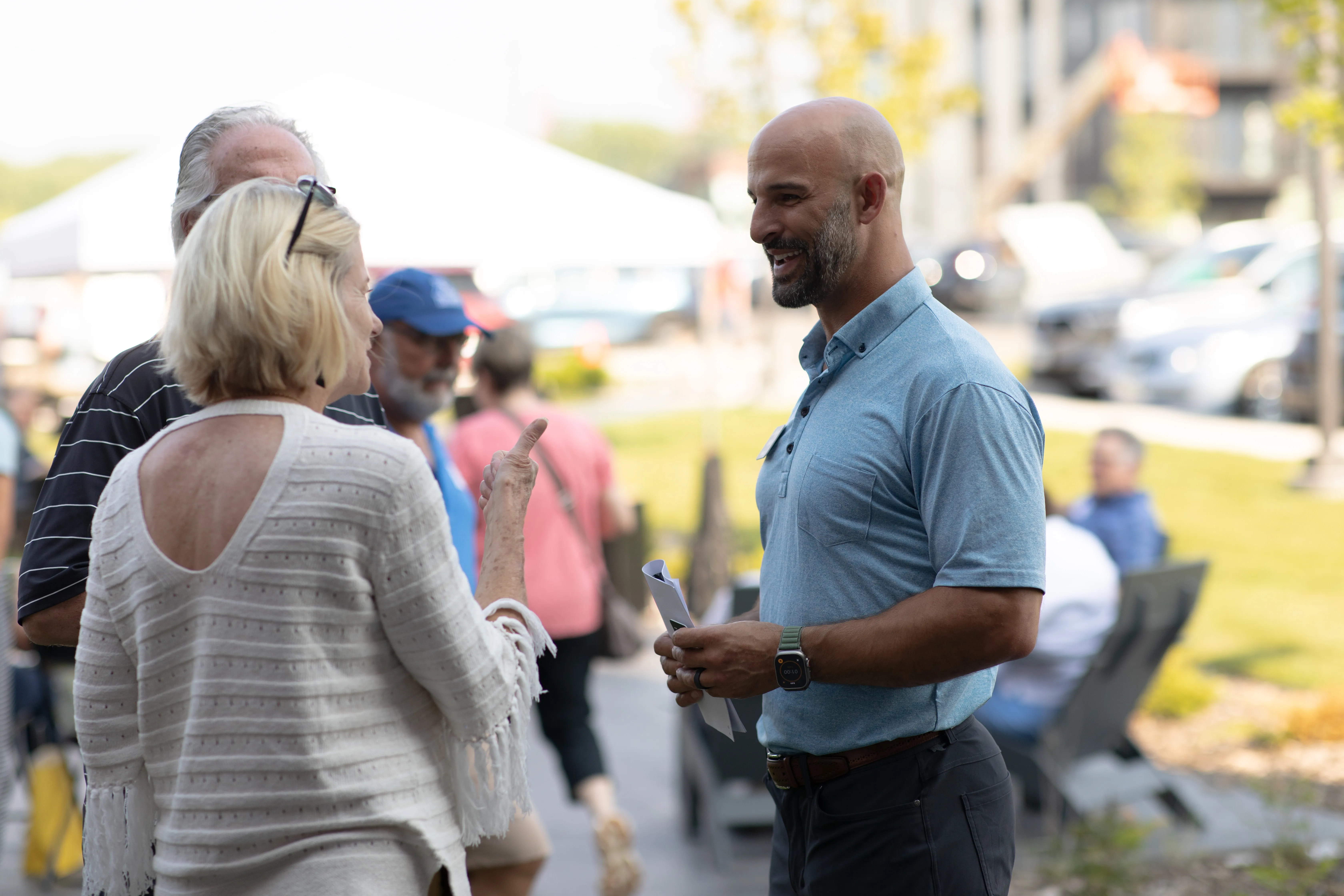 Man in a blue shirt smiling and holding papers while conversing with a woman in a white sweater outdoors.