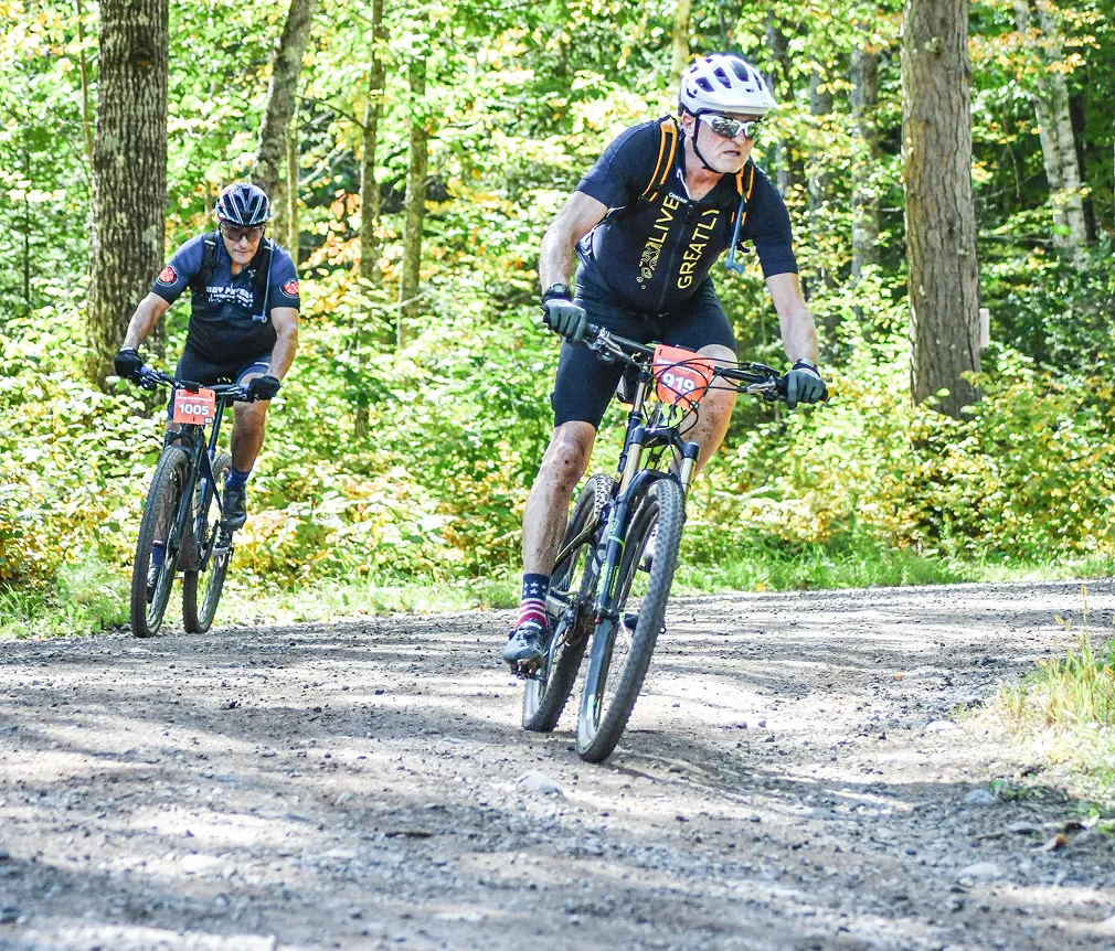 Two cyclists racing on a gravel forest trail surrounded by green trees.