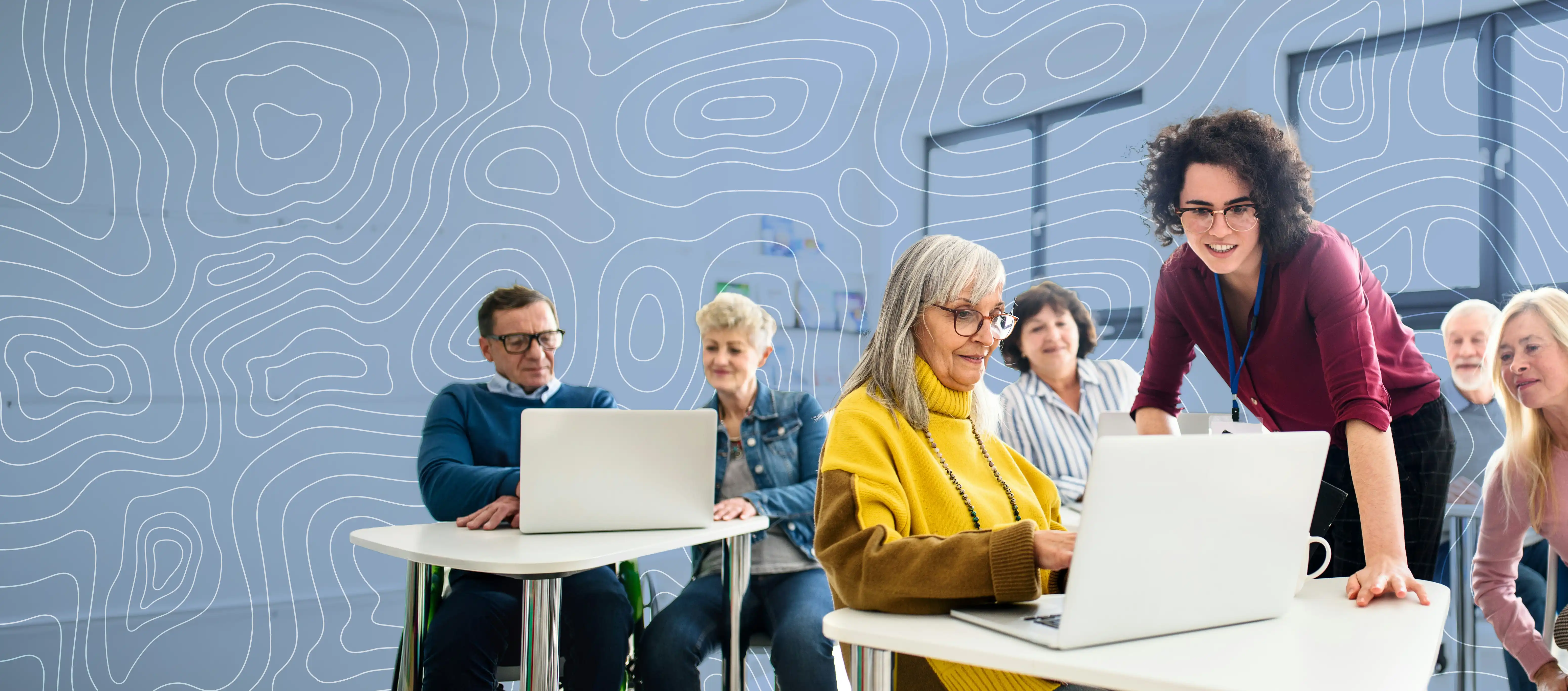 Older adults in a classroom setting using laptops, with a younger woman assisting one of them.