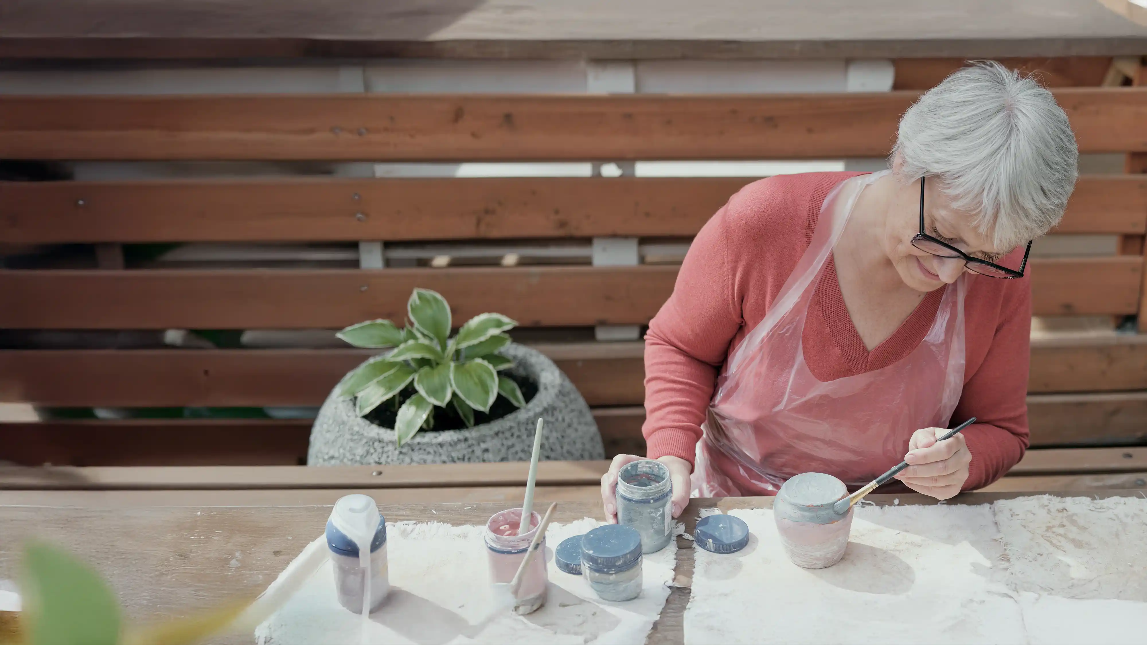 Elderly woman wearing glasses and a pink sweater painting a ceramic pot at a wooden table outdoors.