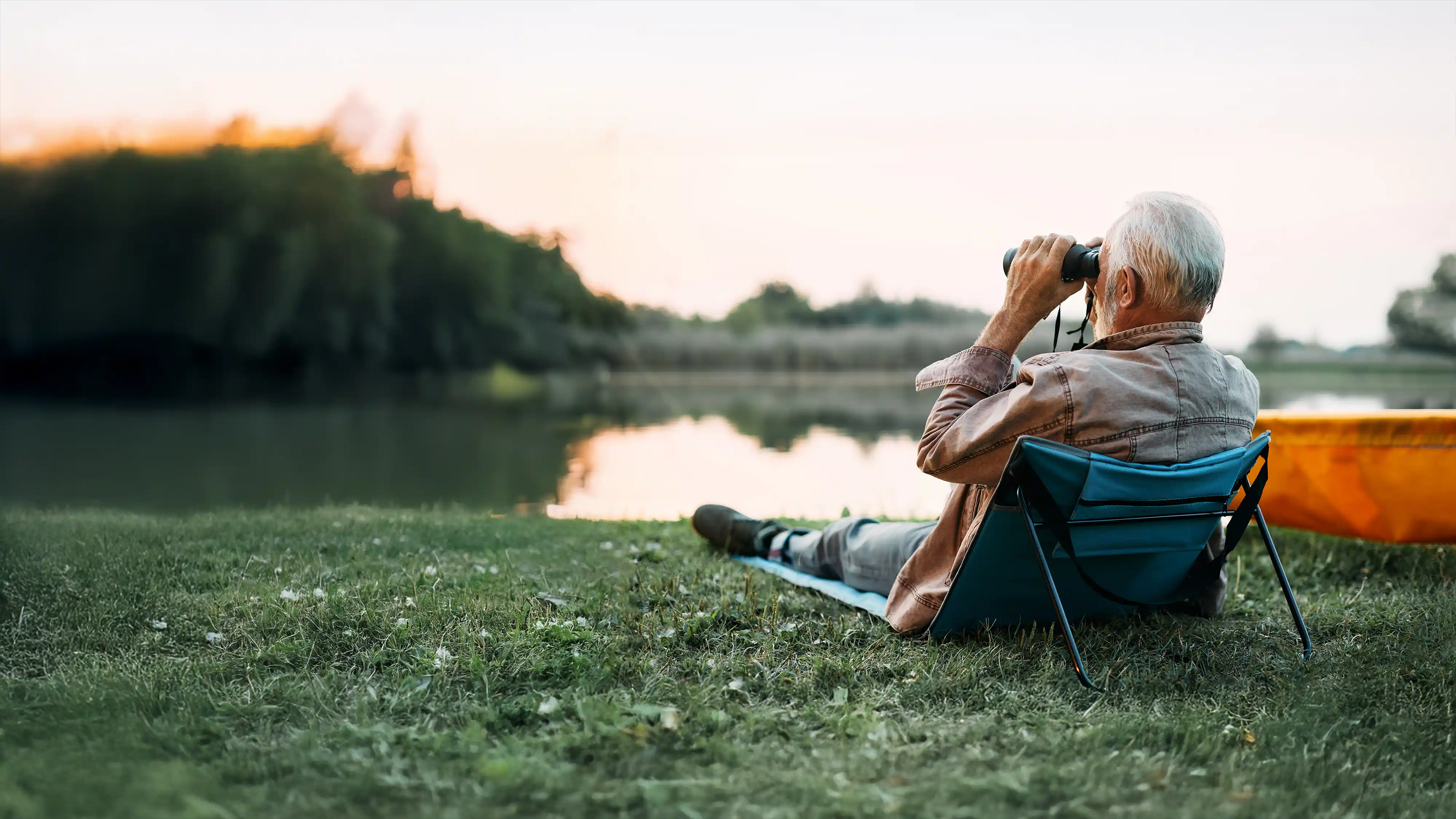 Elderly man sitting on a portable chair by a lake, looking through binoculars at sunset.