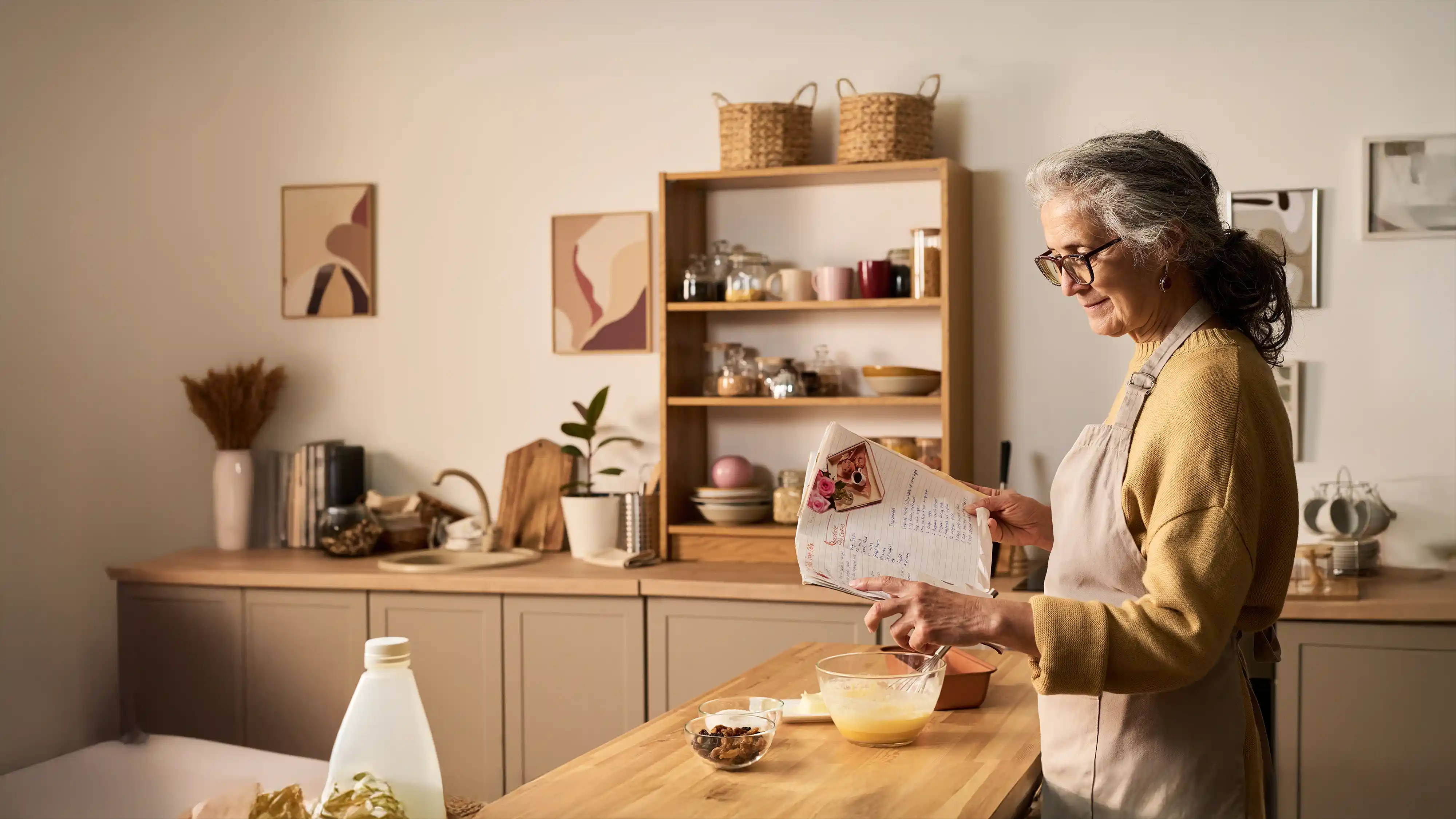 Older woman in glasses and apron reading a recipe in a cozy kitchen while preparing ingredients on the counter.