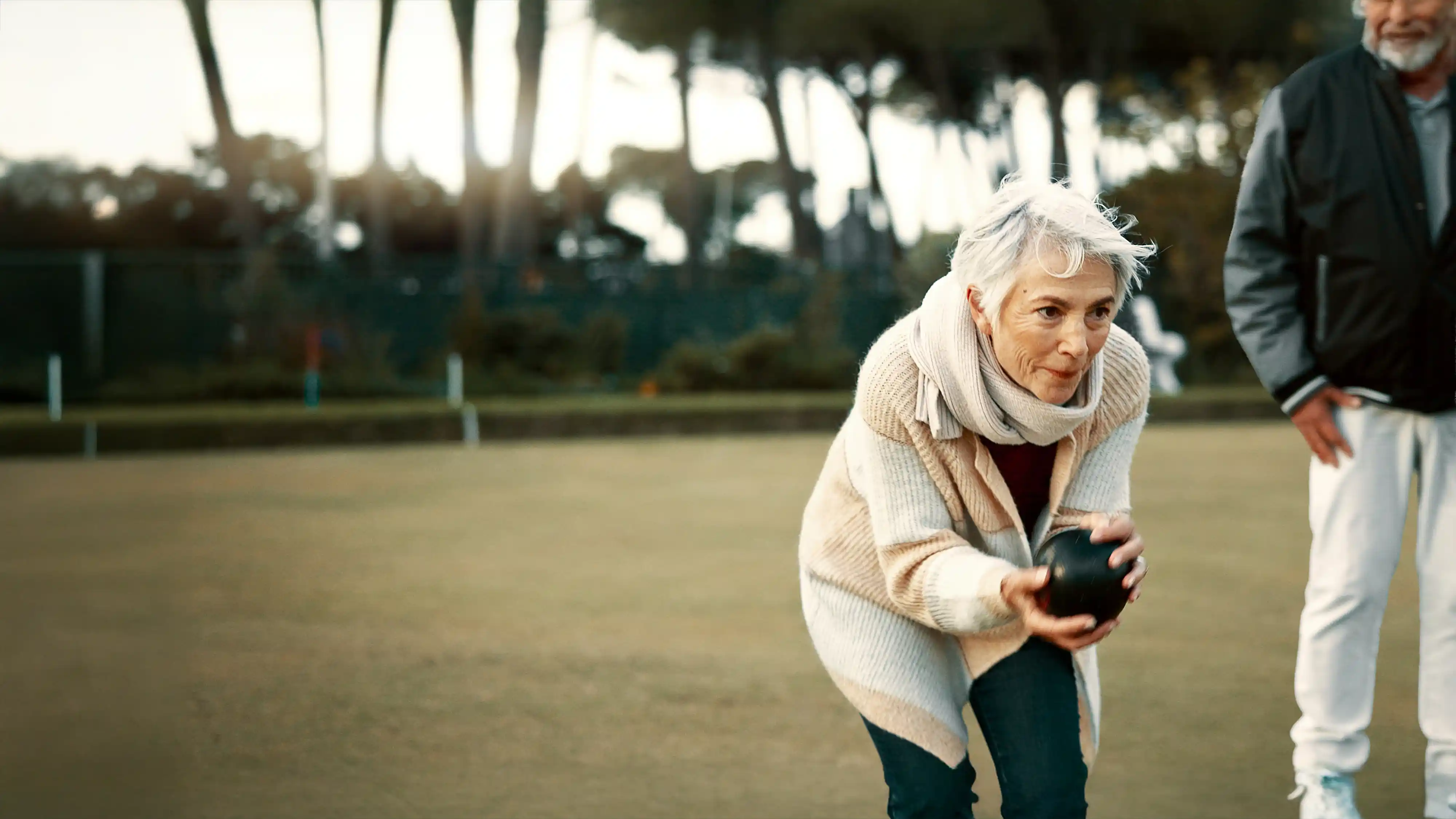 Older woman in a scarf and sweater concentrating as she rolls a lawn bowling ball, with a man watching nearby.