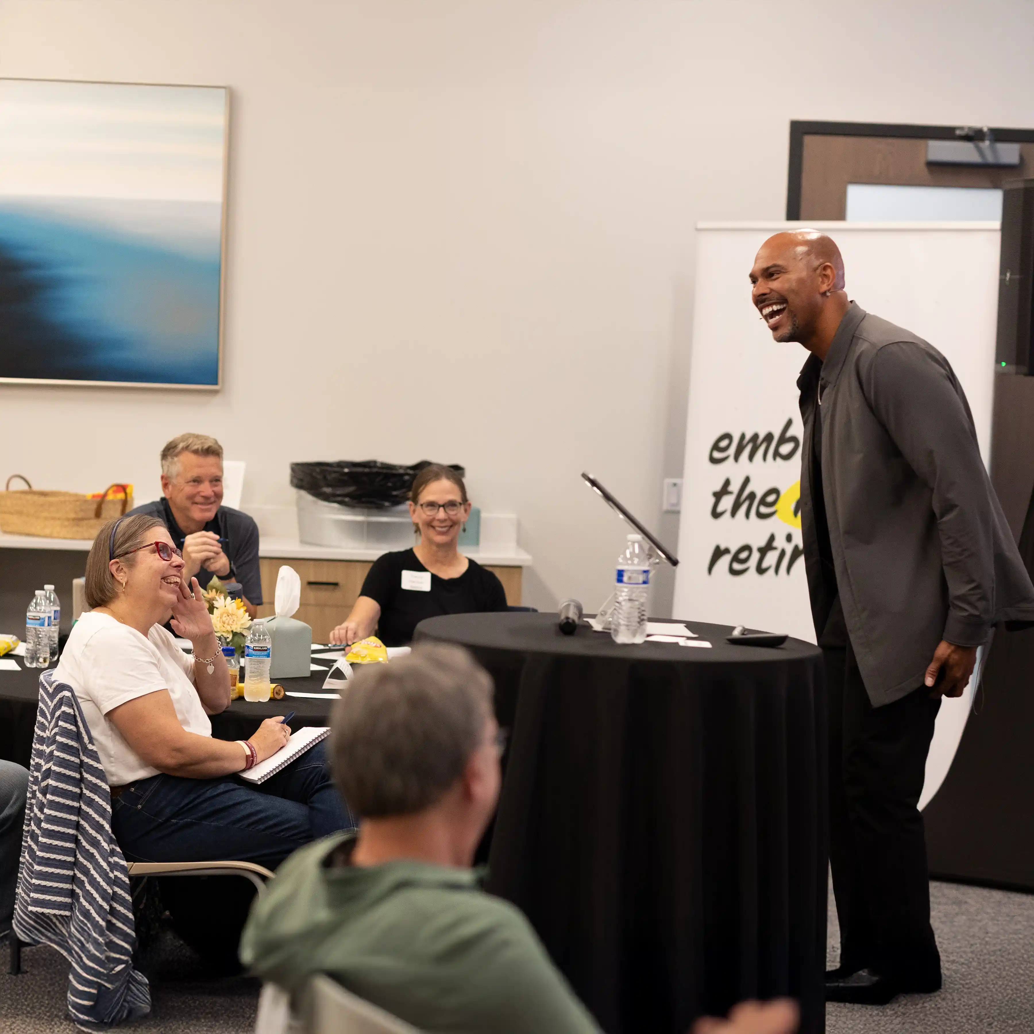 Man giving an engaging presentation to an attentive audience seated around tables in a conference room.