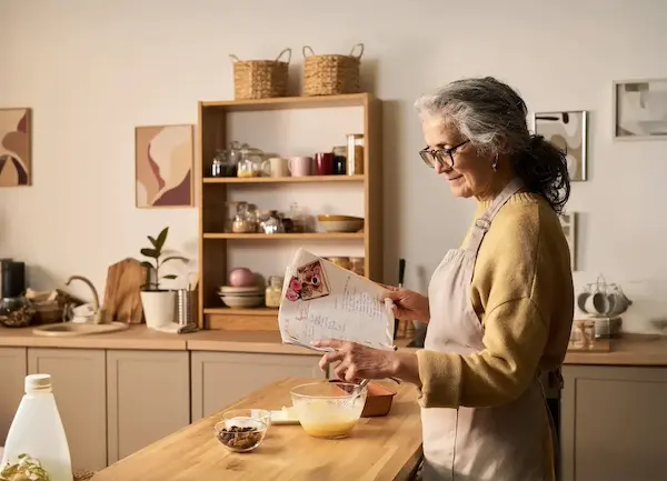 Older woman wearing glasses and apron reading a recipe while mixing batter in a bowl in a warm-toned kitchen.