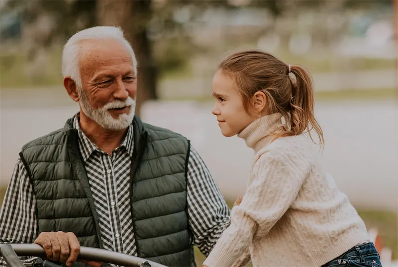 Elderly man with white hair and beard smiling at a young girl wearing a cream sweater outside.
