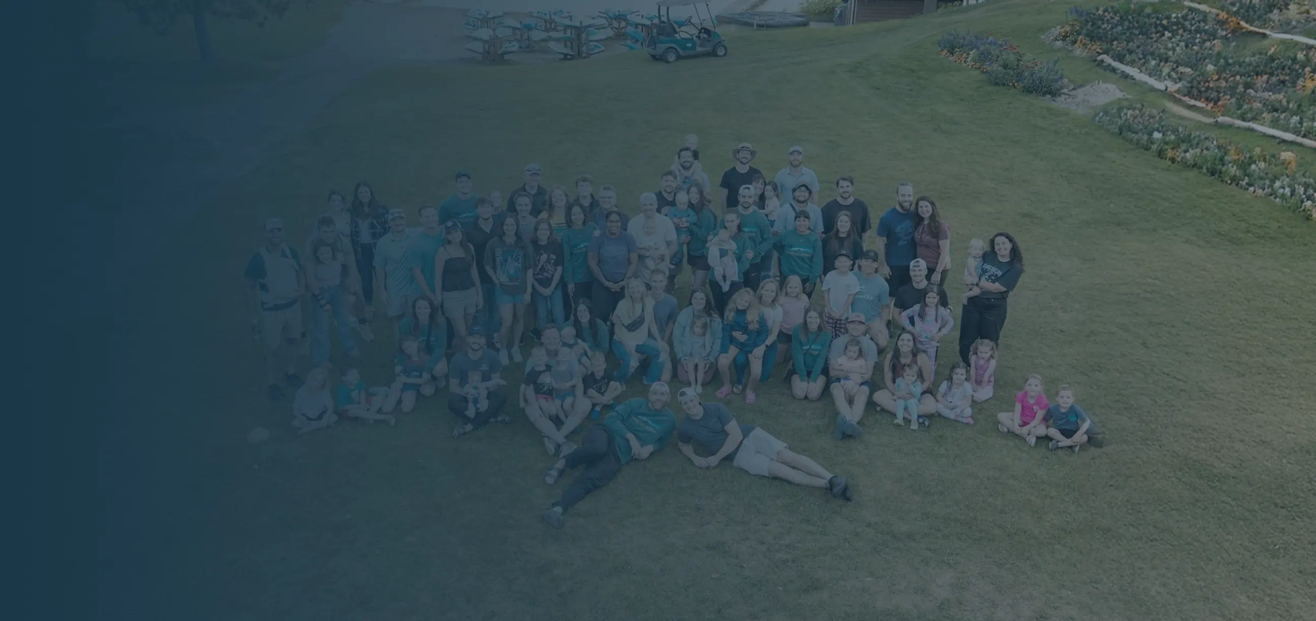 Large group of people, including adults and children, gathered together sitting and standing on grass outdoors.
