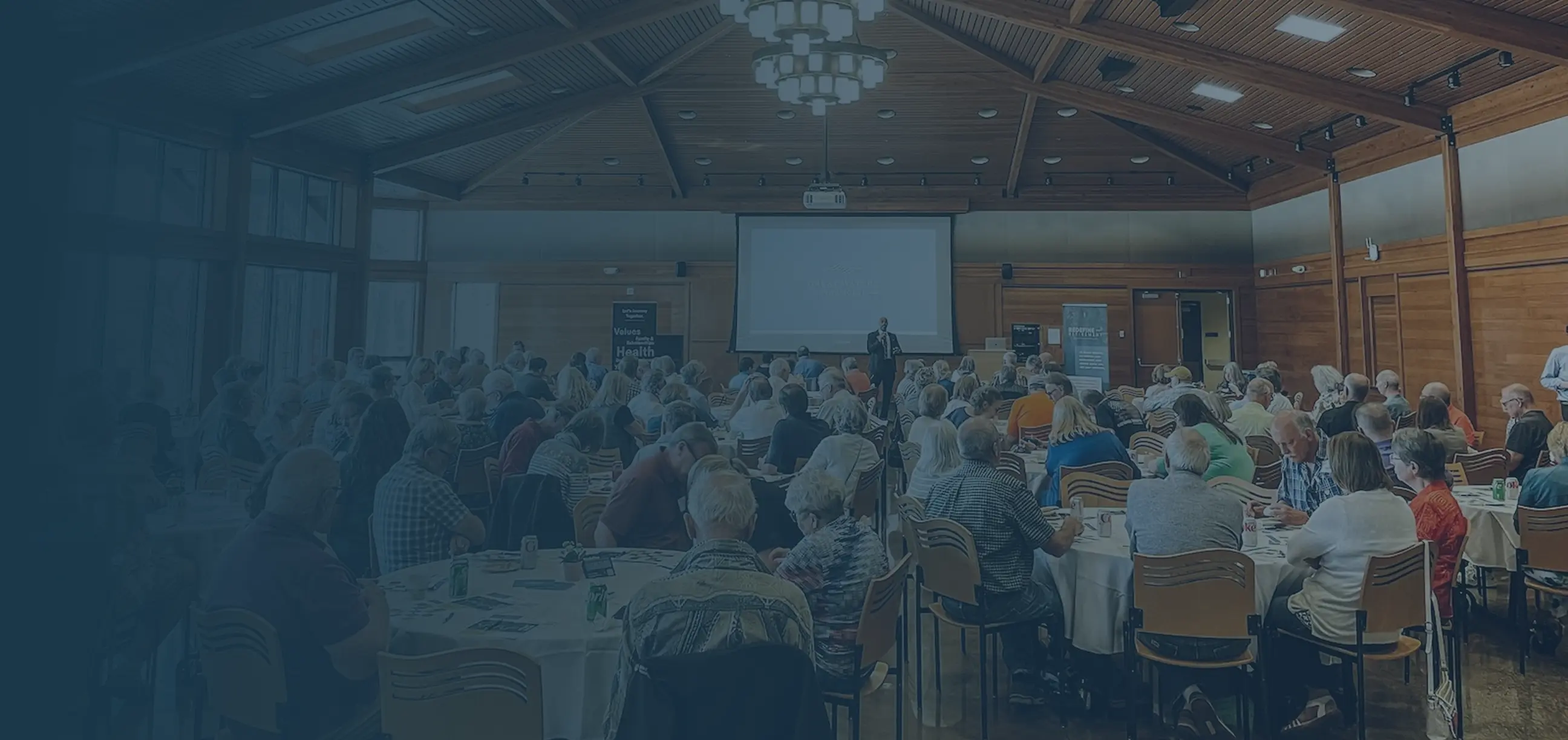 A large group of people seated at round tables in a conference room listening to a speaker presenting in front of a projection screen.