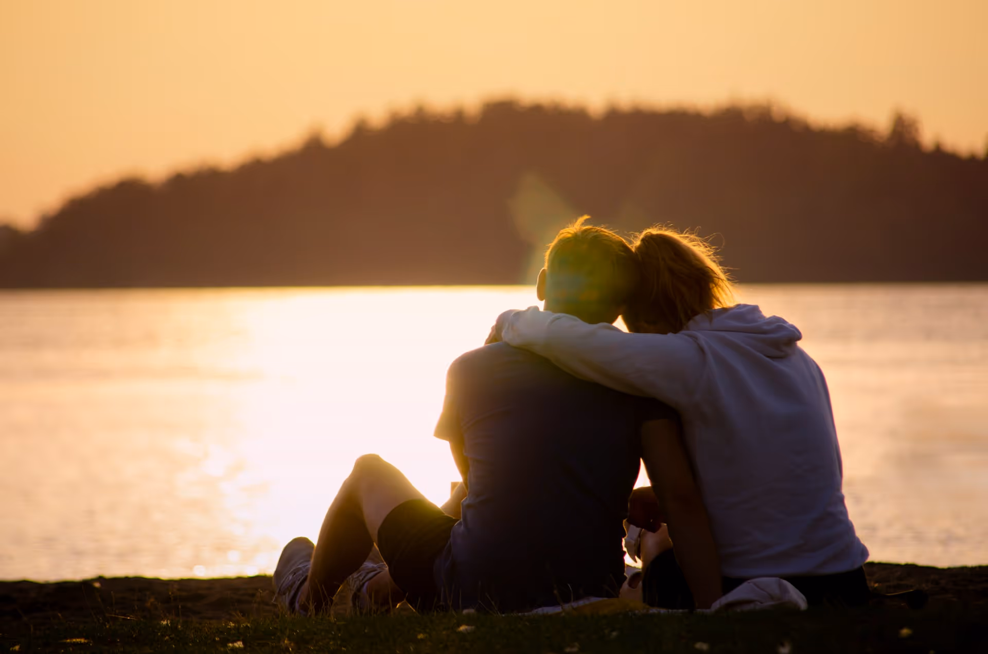Couple sitting on grass by a lake, embracing while watching the sunset.