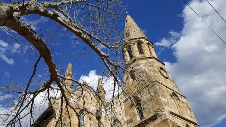 Stone church tower with a pointed steeple framed by leafless tree branches against a blue sky with clouds.