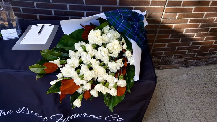 Bouquet of white carnations with green leaves wrapped in blue plaid paper on a funeral company table with a clipboard and pen.