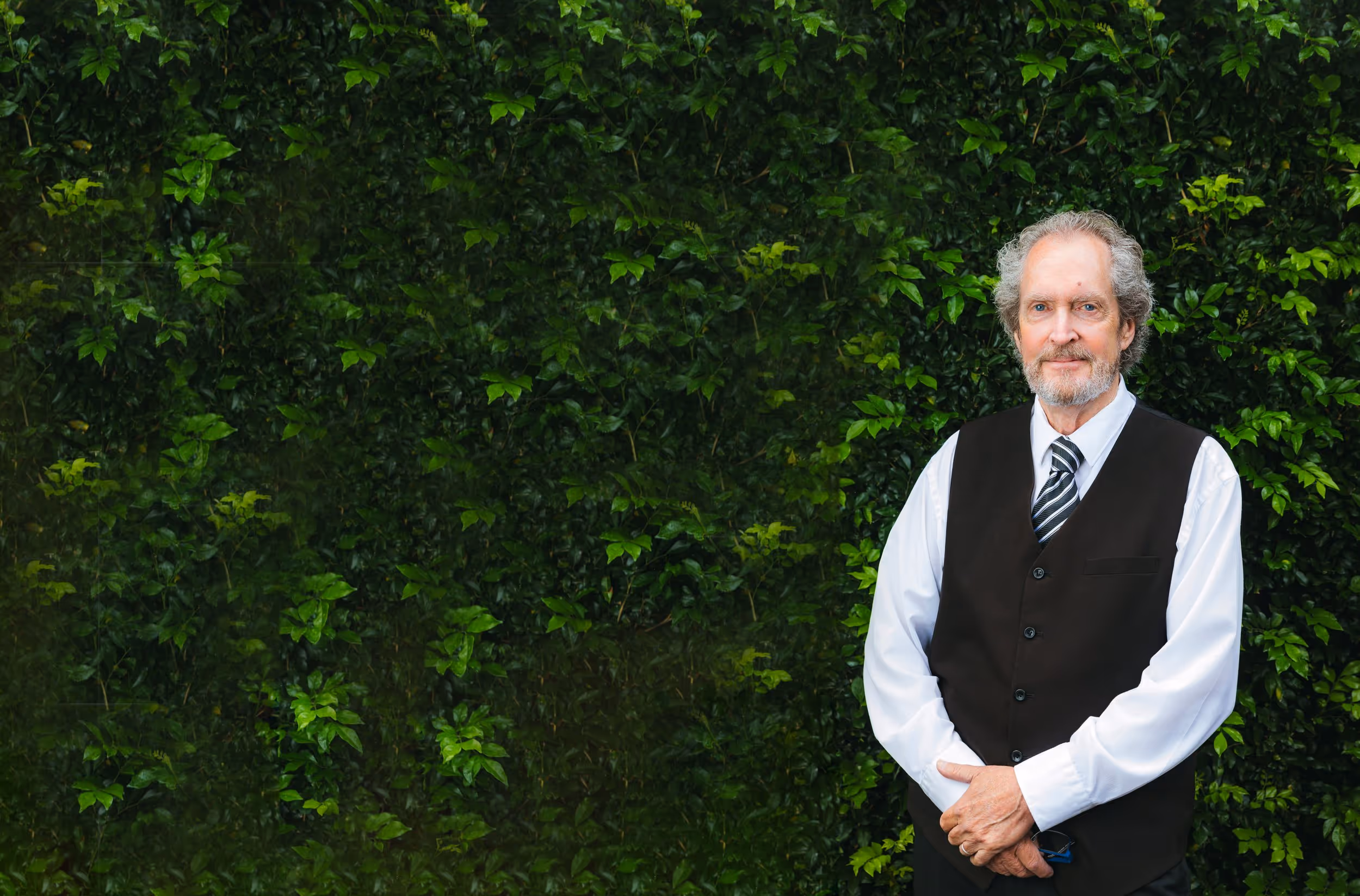 Man with beard wearing a white shirt, striped tie, and black vest standing against a green leafy background.