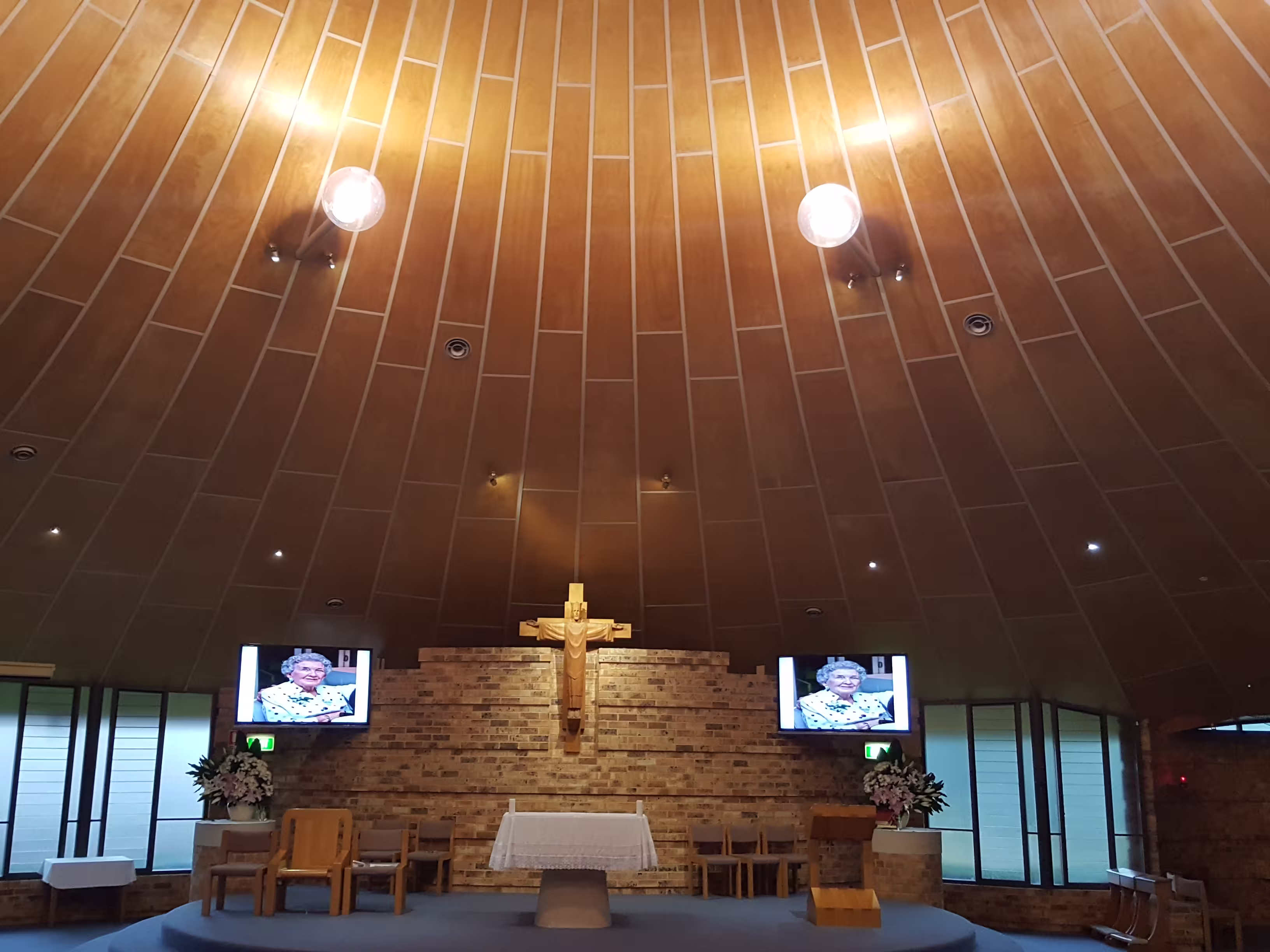 Interior of a church with a wooden crucifix on a brick wall behind an altar, two screens showing an elderly woman, and flowers on either side.
