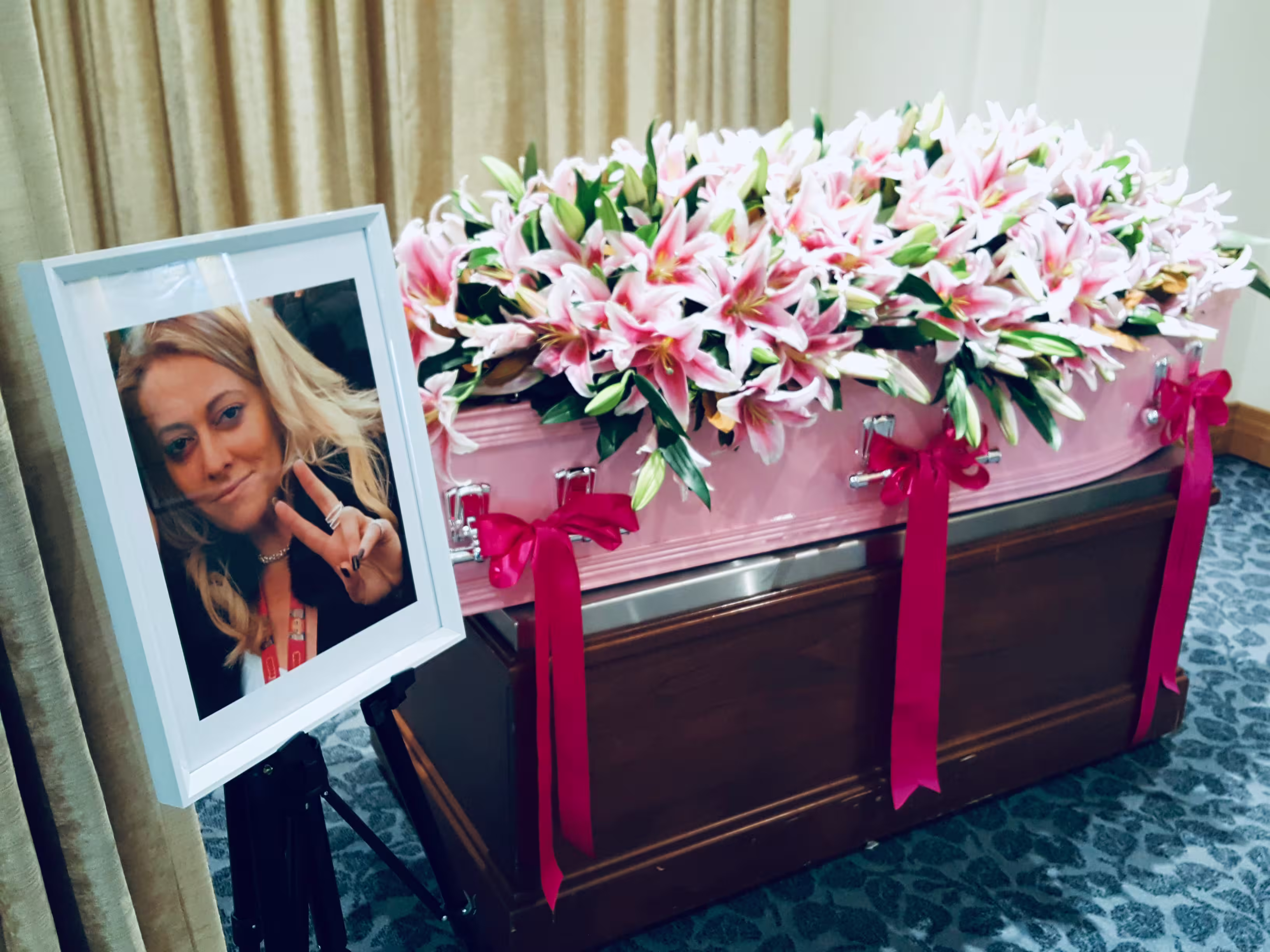 Pink coffin adorned with pink lilies and red ribbons next to a framed photo of a smiling woman making a peace sign.