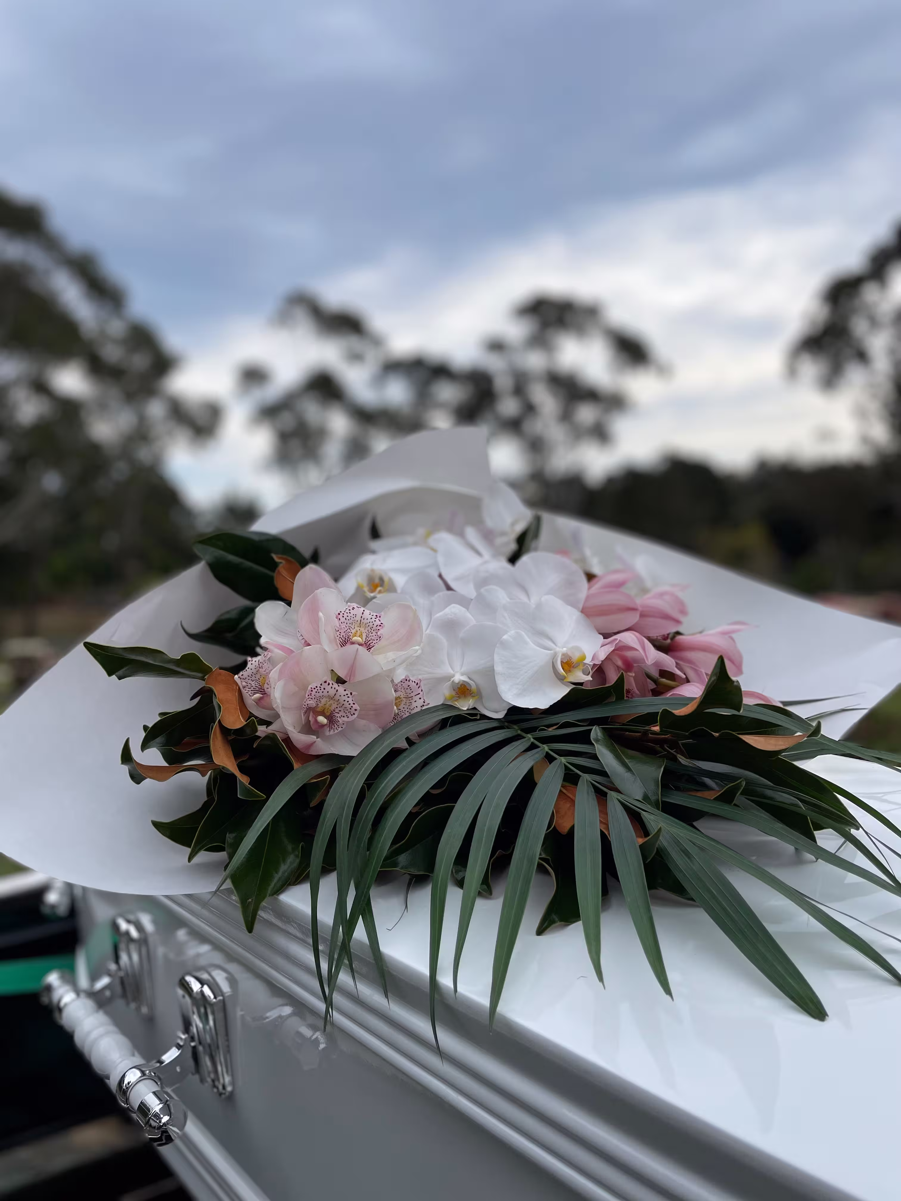 White coffin adorned with a floral arrangement of white and pink orchids and green leaves.