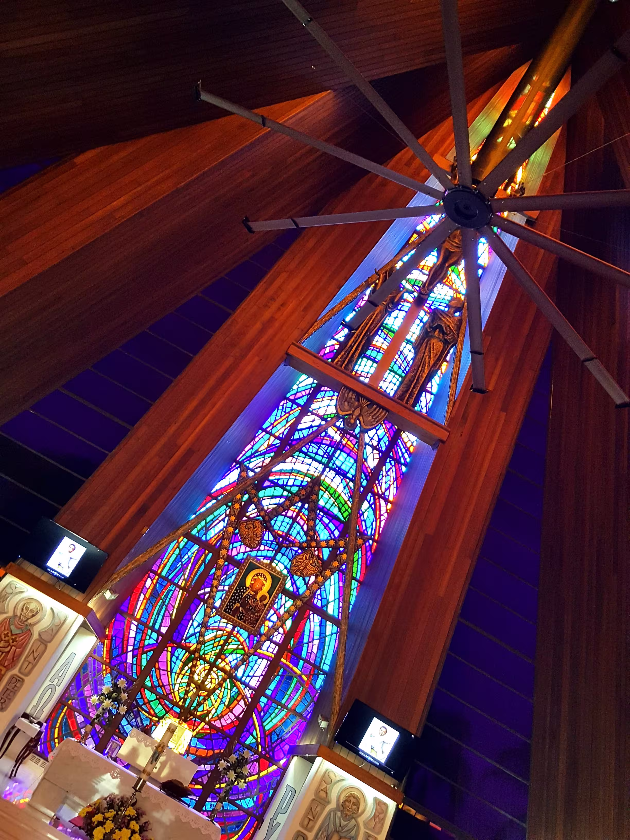 Interior view of a church featuring a large stained glass window behind the altar with religious icons and wooden ceiling beams.
