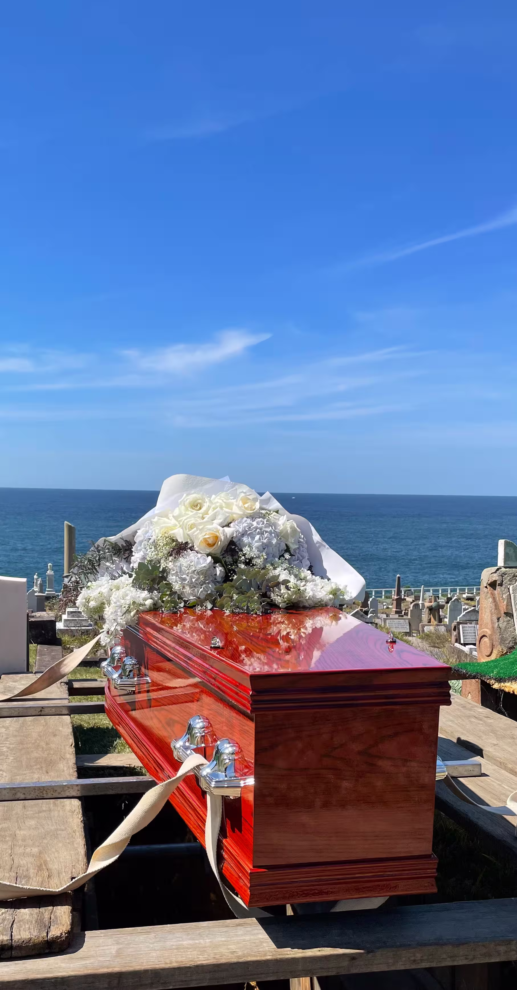Polished wooden coffin with white and pale yellow floral arrangement on top, placed over a burial site with ocean and clear blue sky in the background.