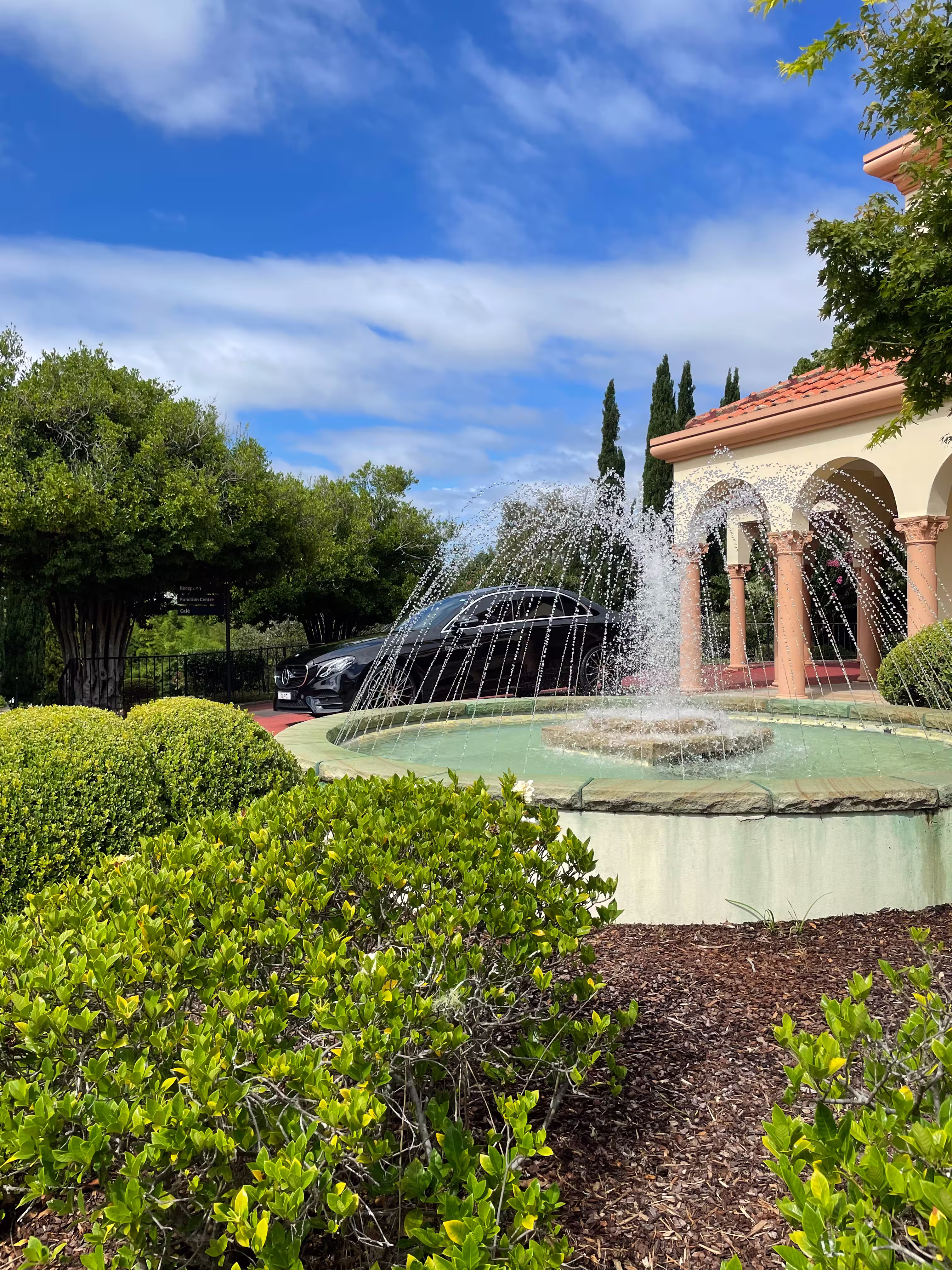 Outdoor fountain with water jets arching around a central spray in front of a Mediterranean-style building with a terracotta tile roof and green shrubs.
