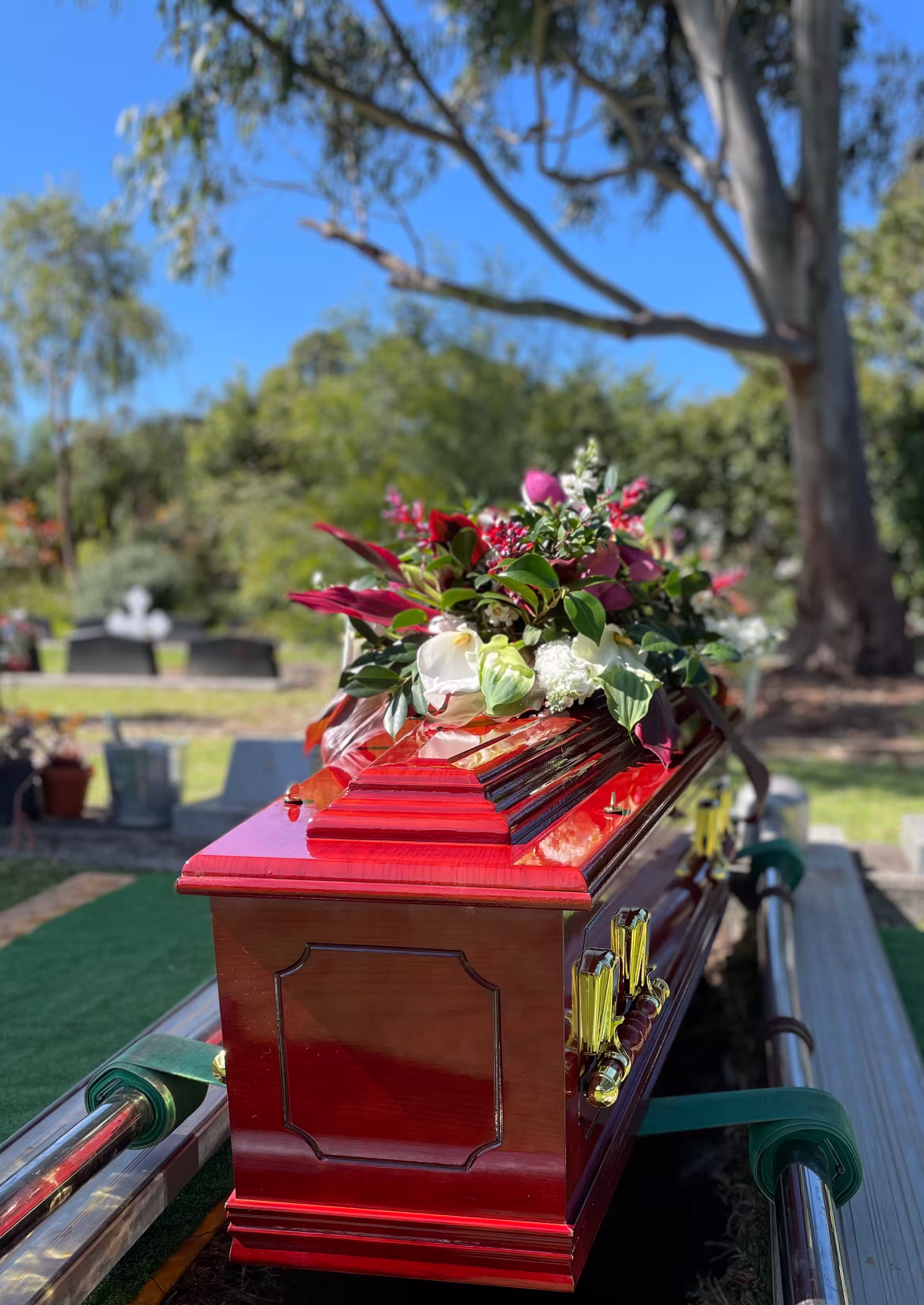Glossy red coffin adorned with a floral arrangement of white and pink flowers in an outdoor cemetery setting.