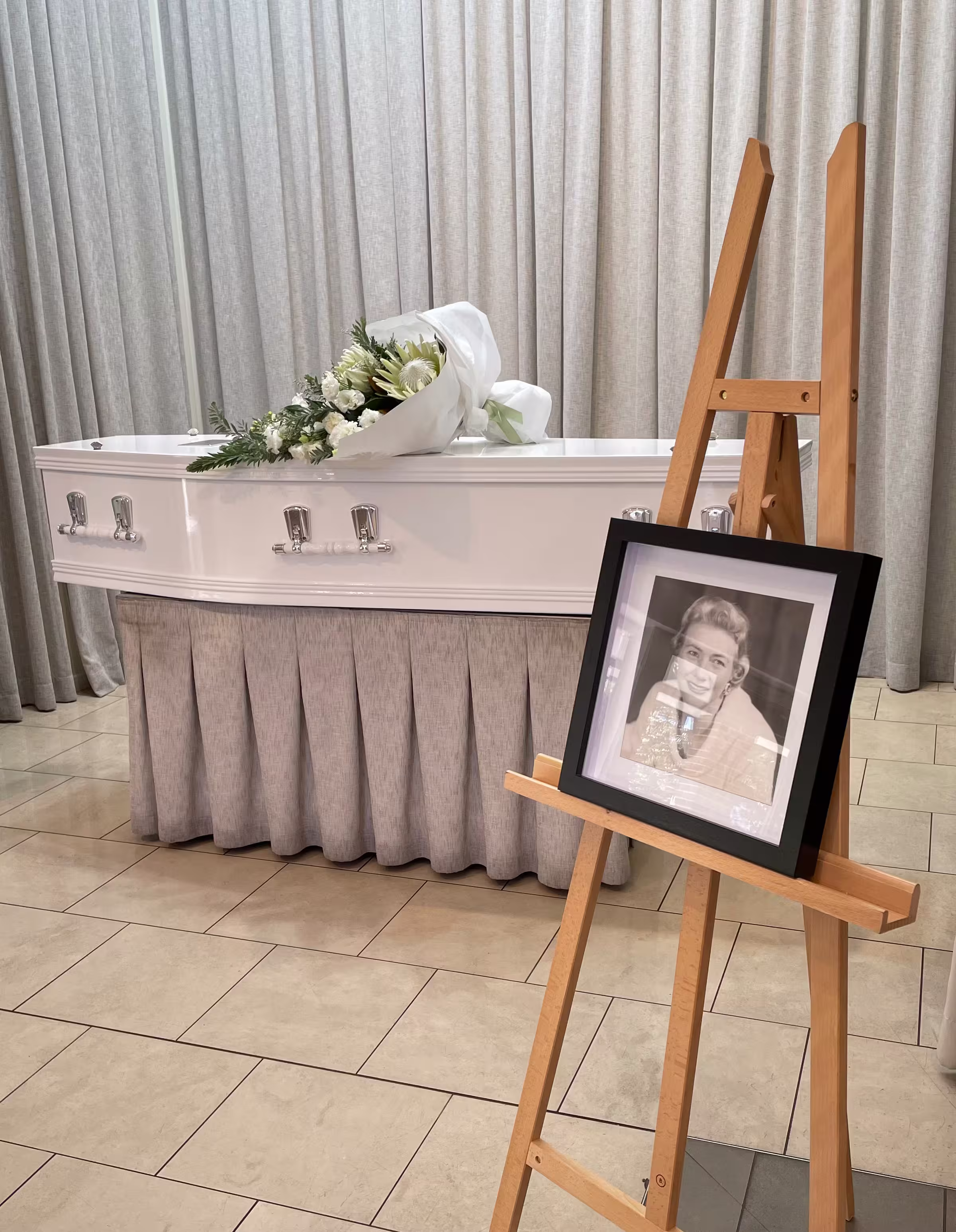 White coffin with a bouquet of white flowers on top, next to an easel holding a black-framed portrait of a smiling woman, in a room with beige curtains and tiled floor.