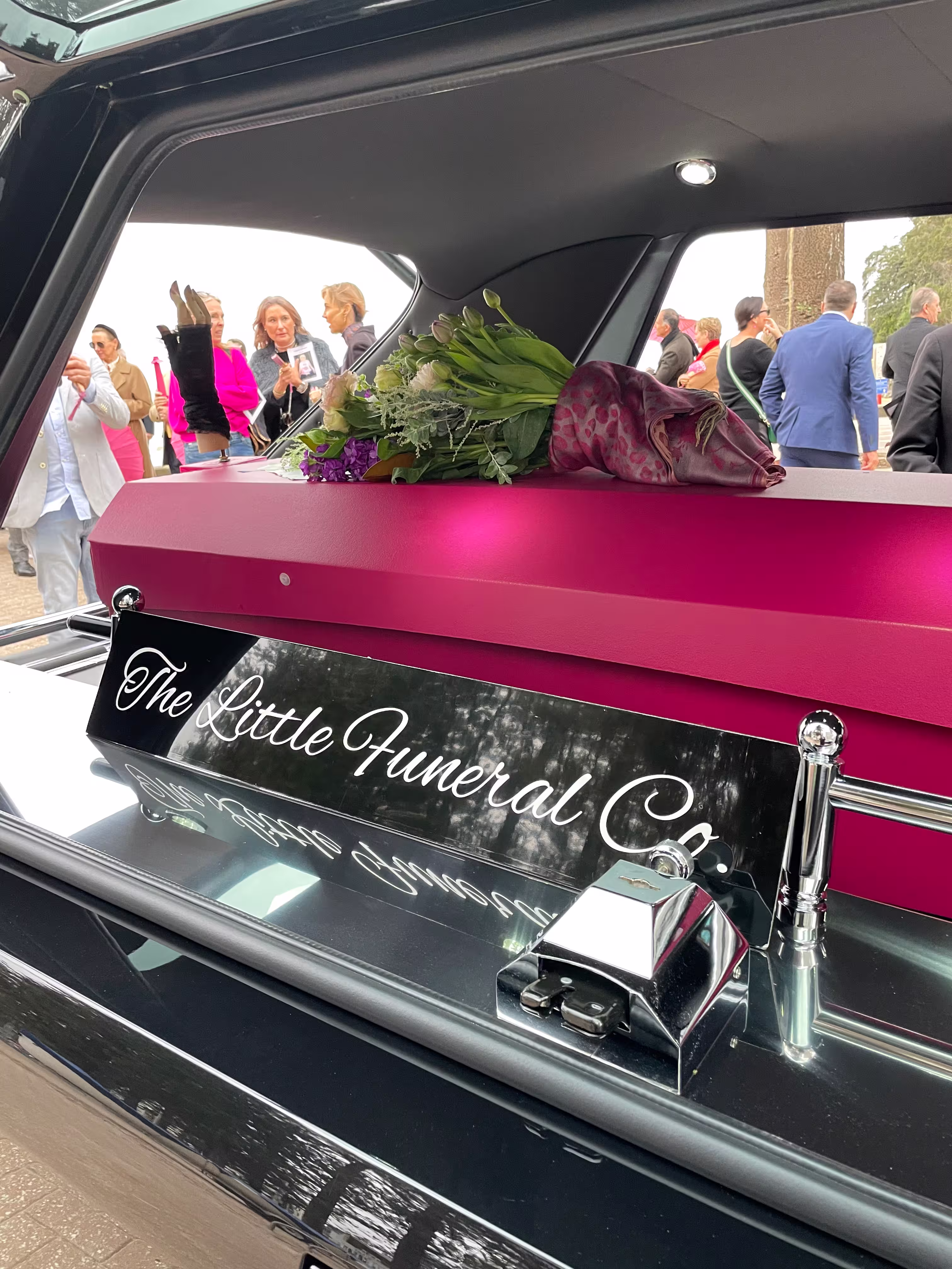 Pink coffin with flowers on top inside a hearse with a sign reading 'The Little Funeral Co.' and people standing outside.