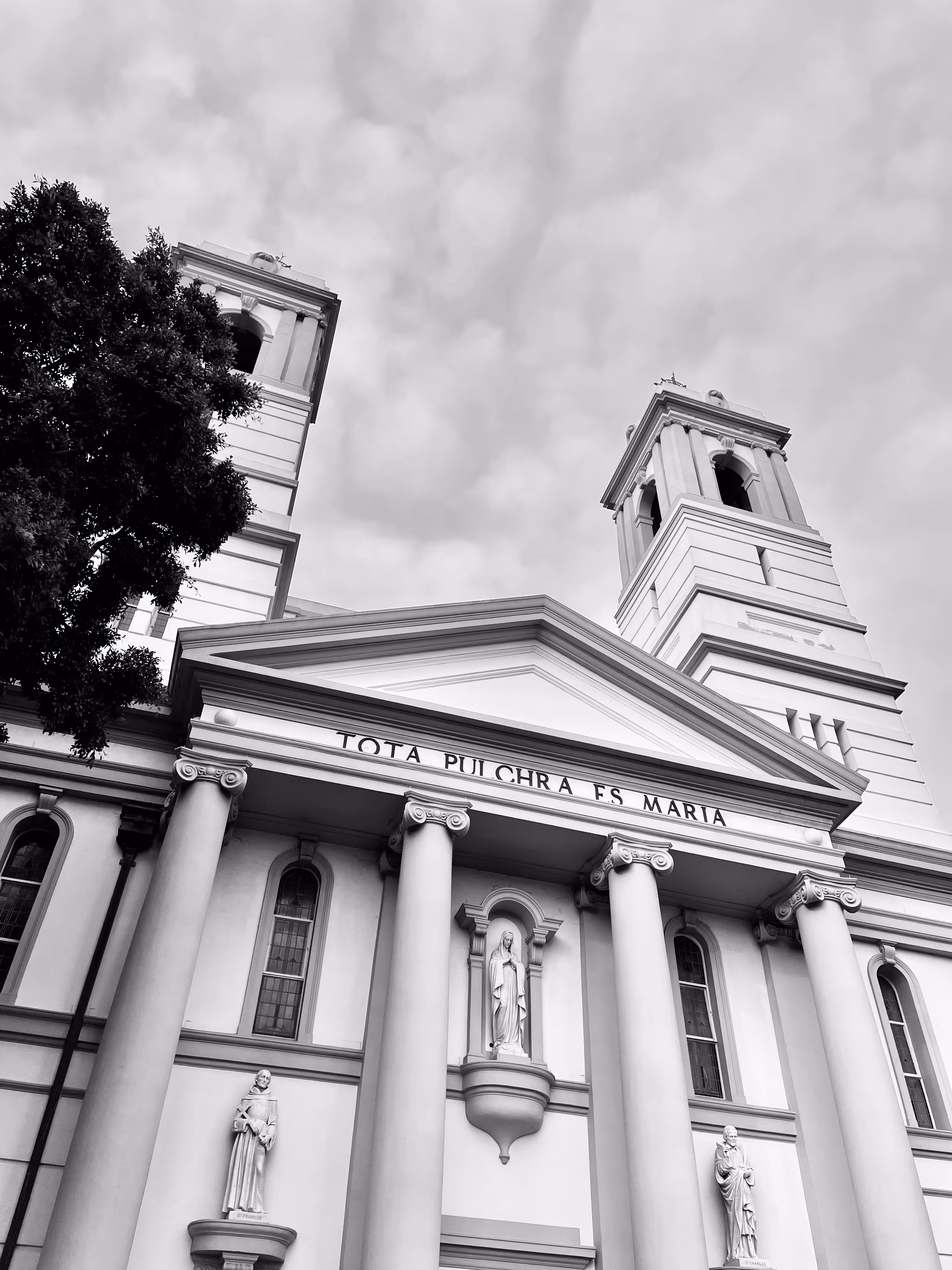 Front view of a classical church facade with tall columns, statues, and two bell towers under a cloudy sky.