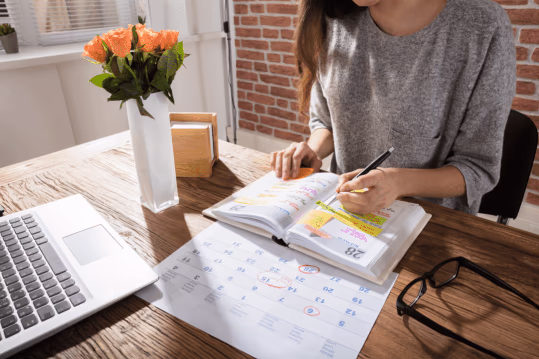 woman in sweater writing in planner on desk with flowers and computer