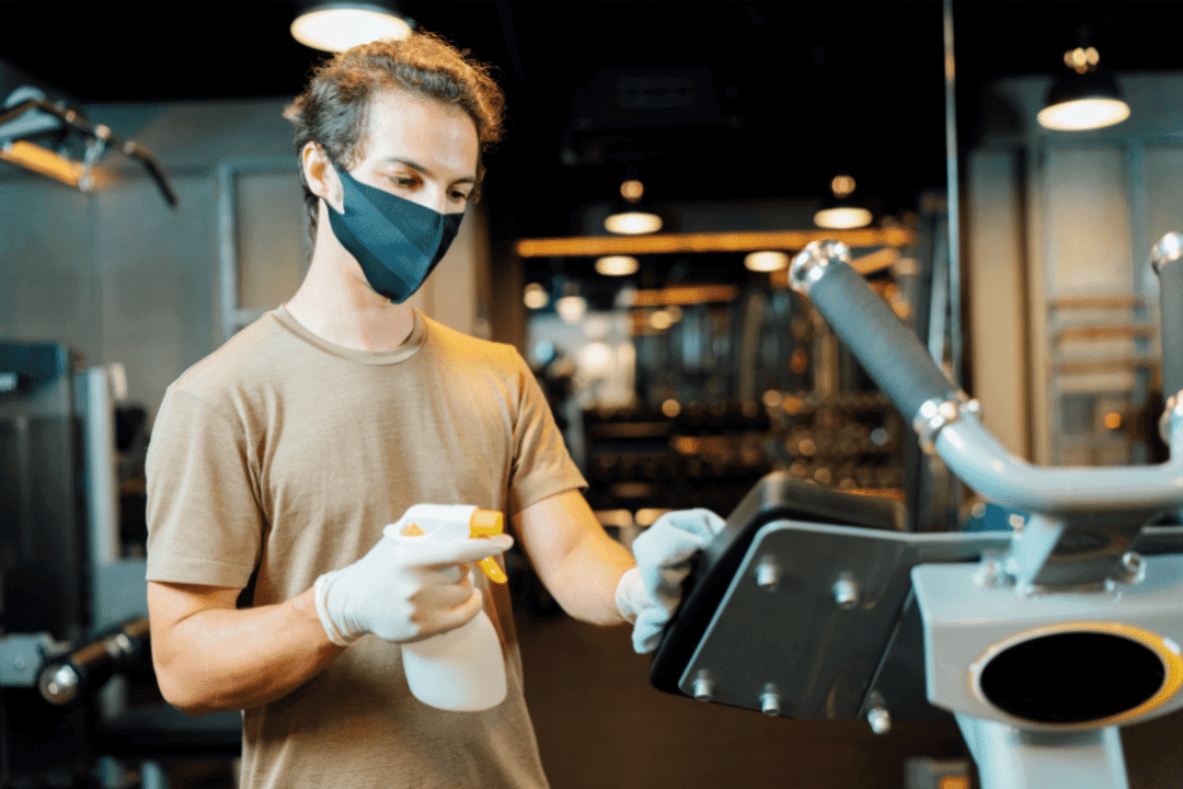 young man wearing mask sanitizing gym equipment with spray bottle