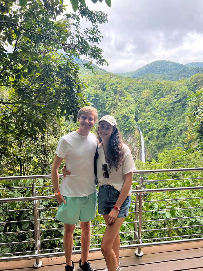 A man and woman smile in front of a waterfall in the distance.
