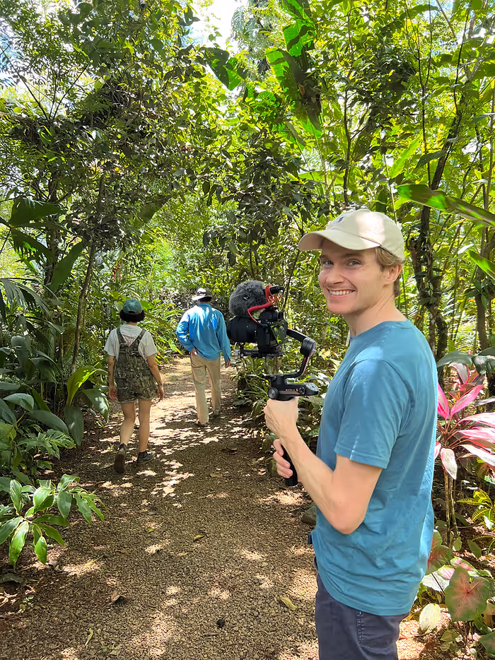 A man with a camera smiles on a dirt trail surrounded by jungle