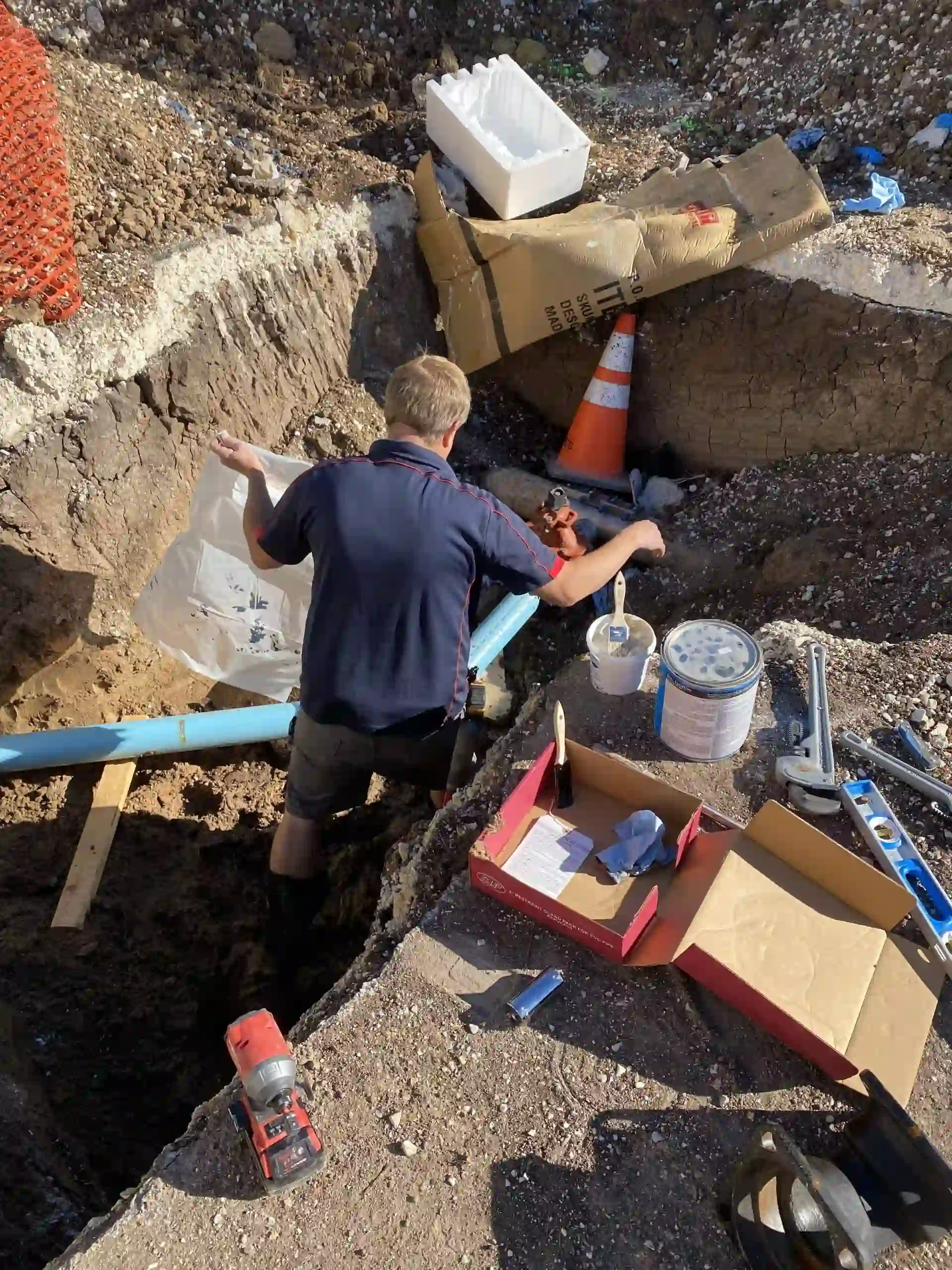 Man in navy shirt working in a dug trench with plumbing pipes and various tools scattered around.