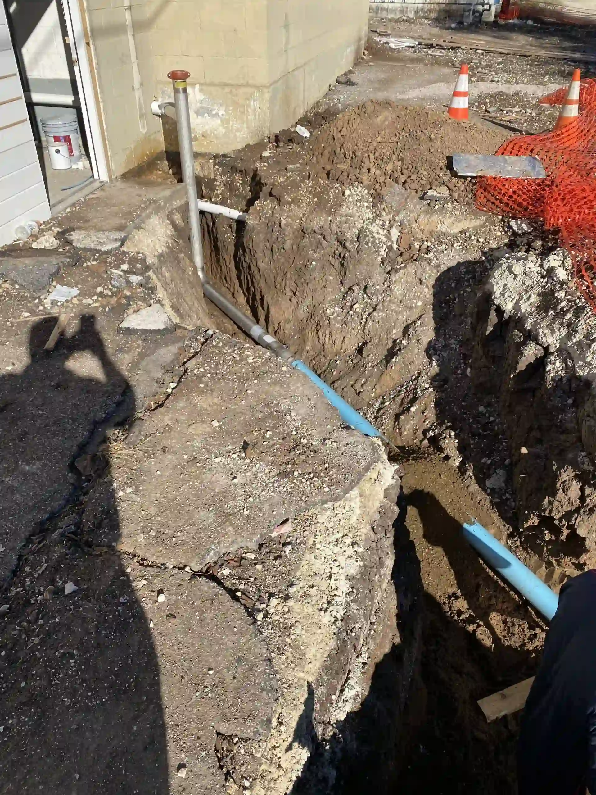 Construction site with a deep trench dug alongside a building foundation, exposing earth and a blue utility pipe, with orange safety cones and orange mesh fencing nearby.