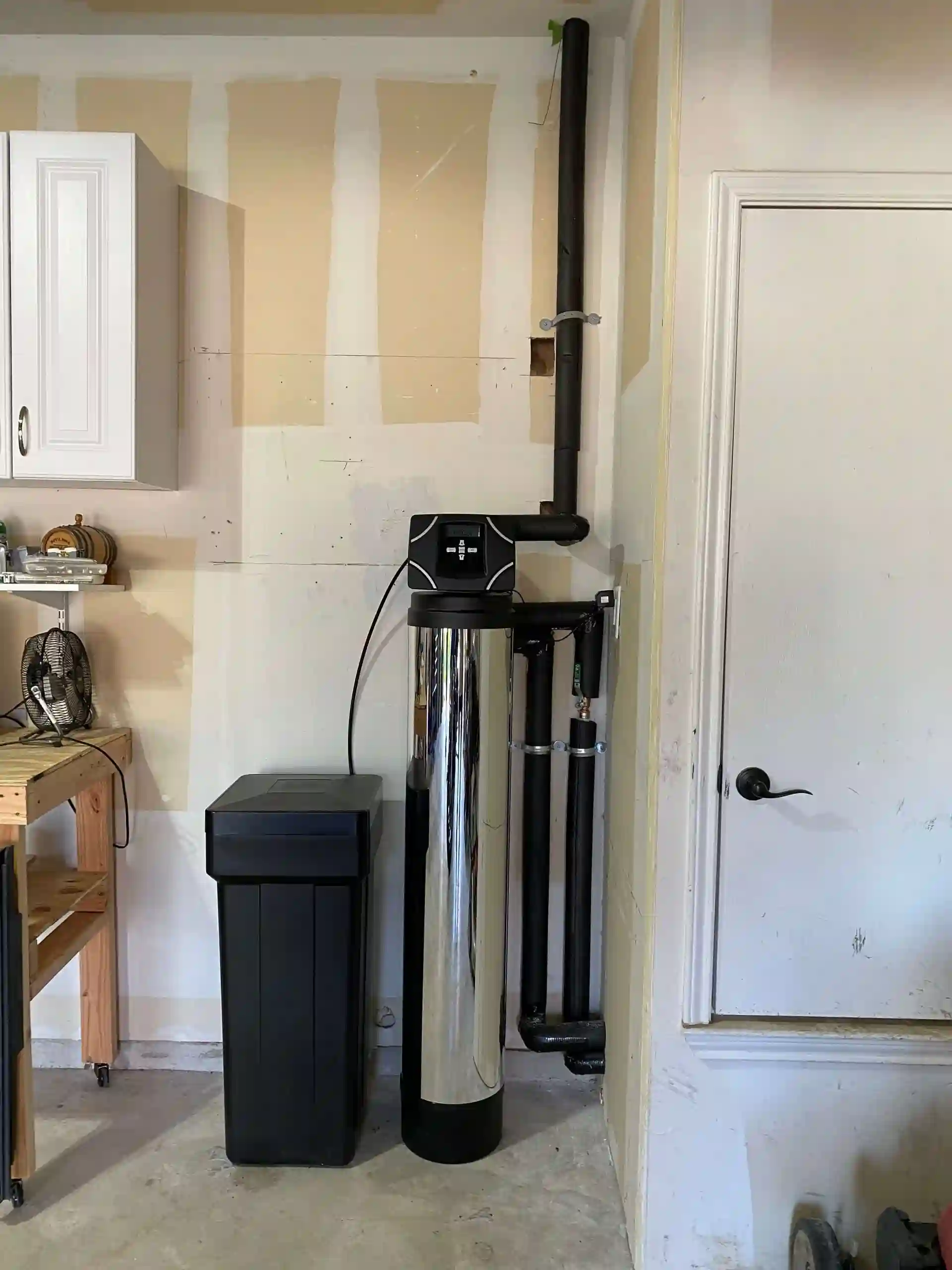 Water softener system installed in a garage corner with black pipes and control panel.