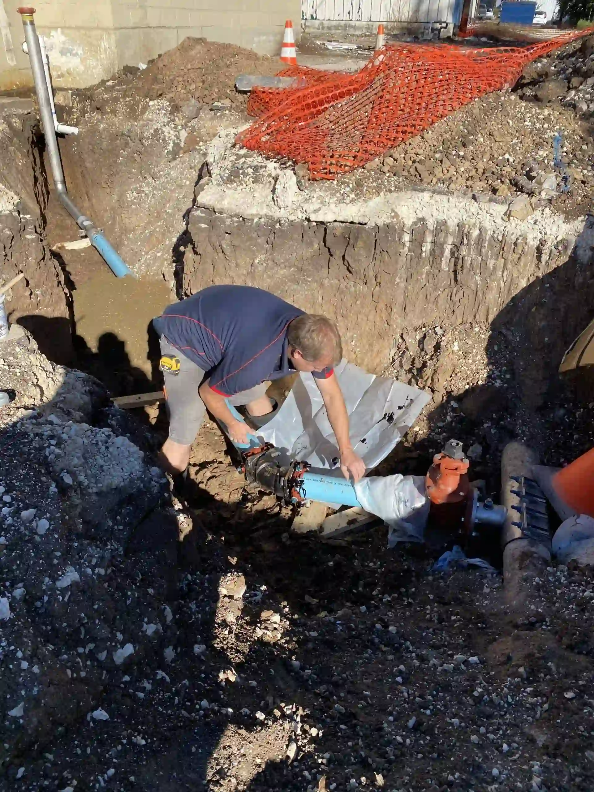 Worker installing or repairing blue underground pipes in a trench with orange safety fencing nearby.