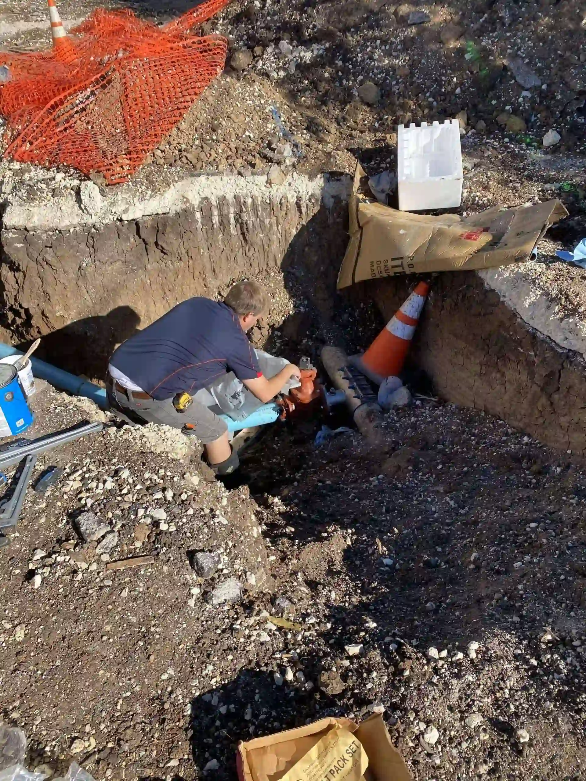 Worker in a trench installing or repairing a blue pipe connector with orange valve, surrounded by dirt, construction cones, and equipment.