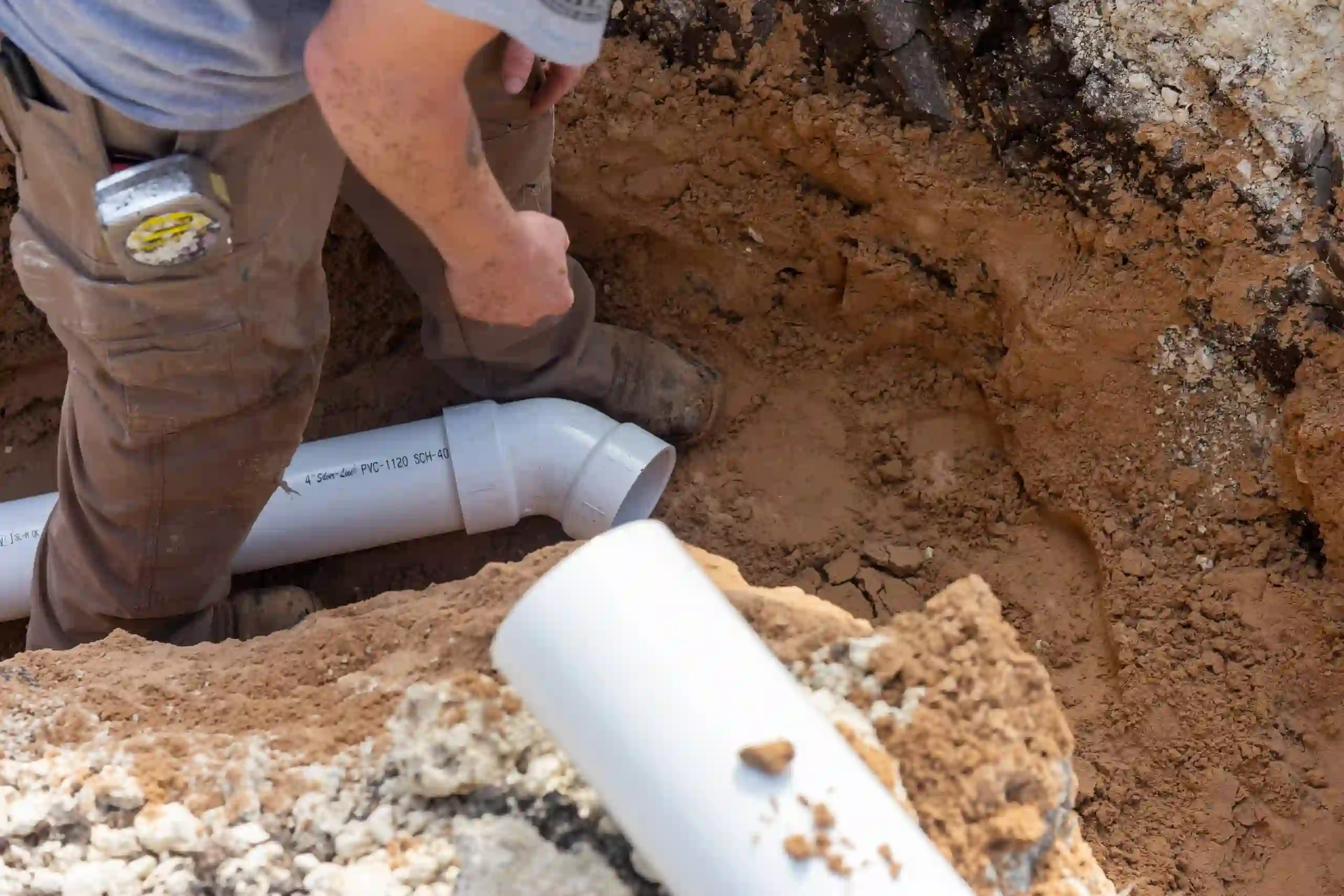 Worker installing white PVC pipes in a dirt trench for drain repair.