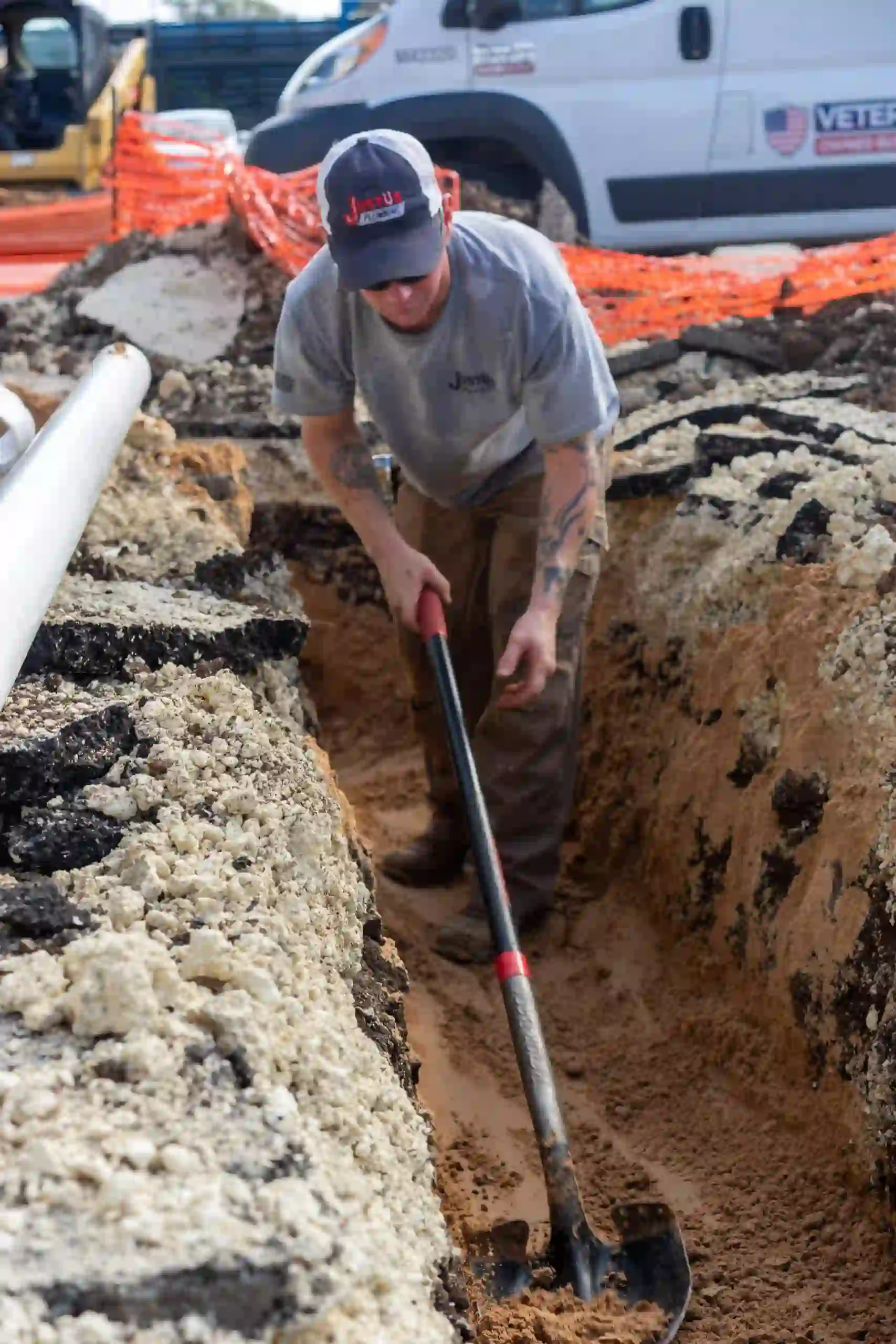 Construction worker digging a trench with a shovel near white pipes and an orange safety fence.