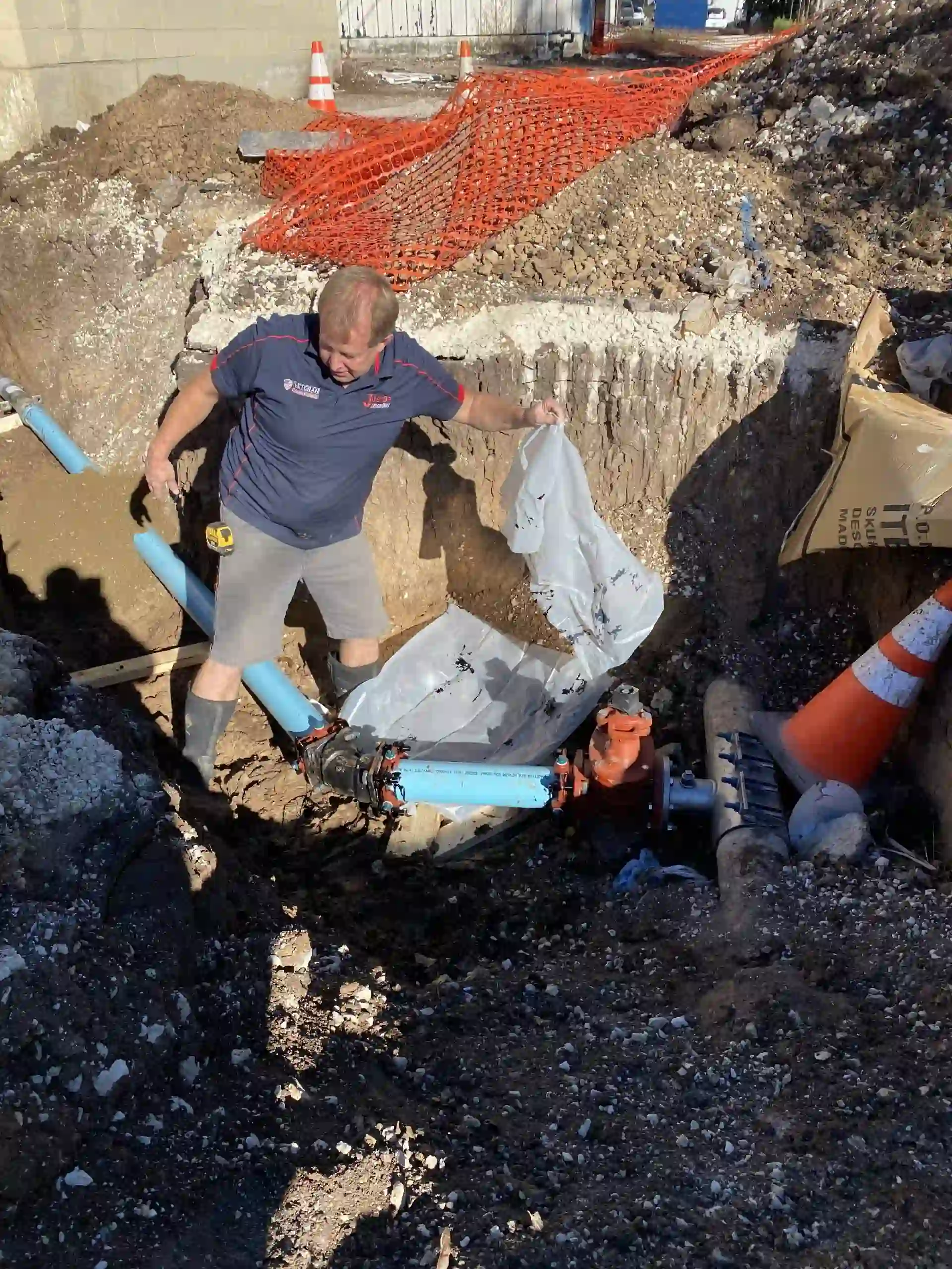 Worker in a navy shirt and gray shorts installing blue pipes in a construction trench surrounded by orange safety fencing and traffic cones.