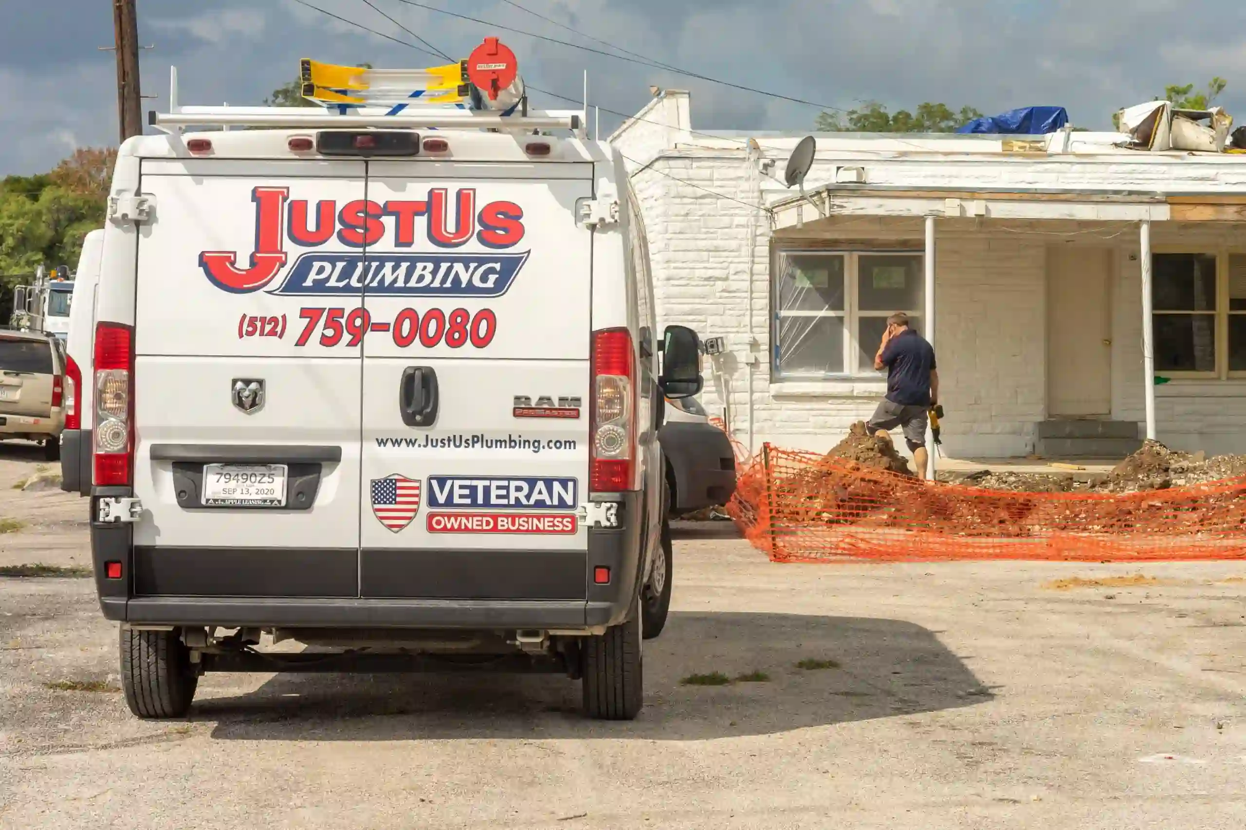 Rear view of a white Just Us Plumbing van parked next to a construction site with an orange safety fence and a man working by a white house.