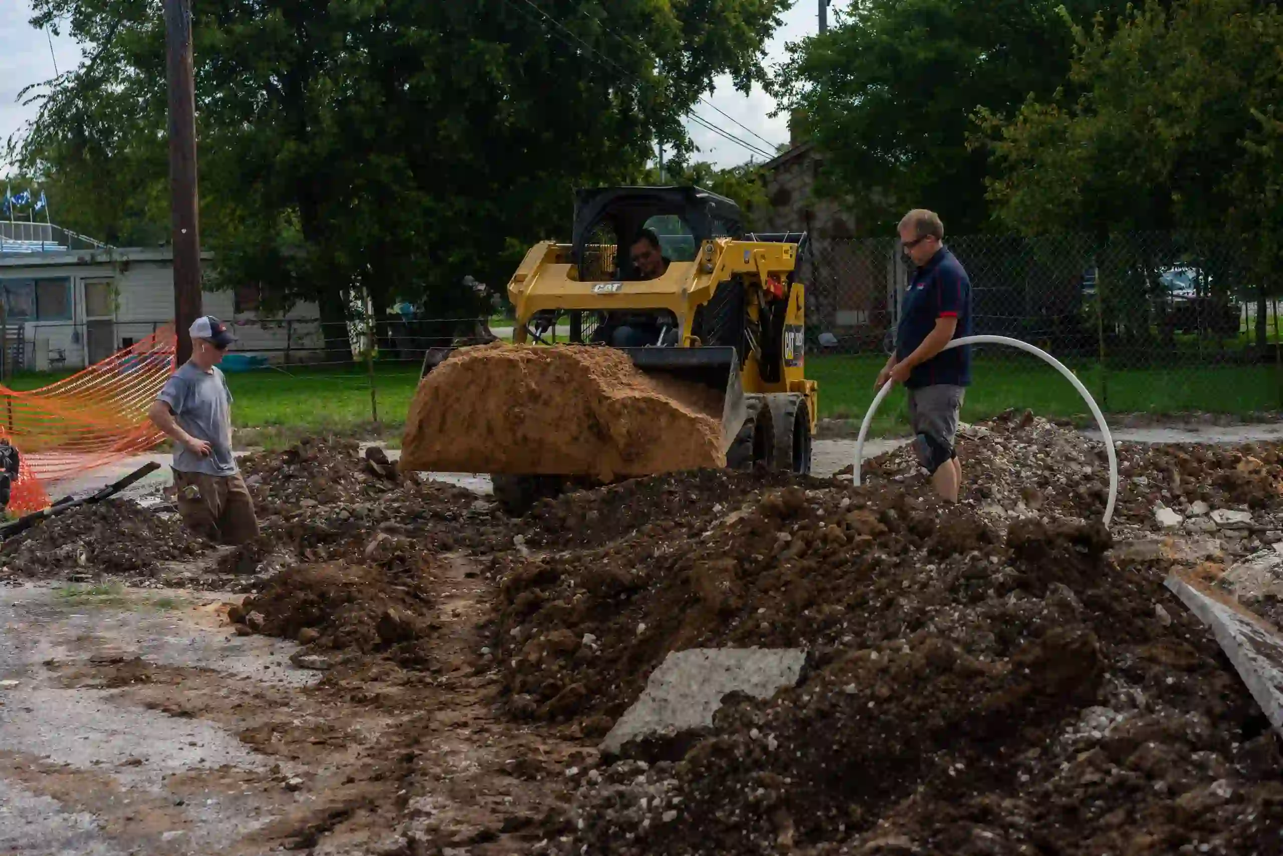 Construction workers operating a yellow skid-steer loader moving dirt at an outdoor site with piles of soil and one worker holding a large white pipe.