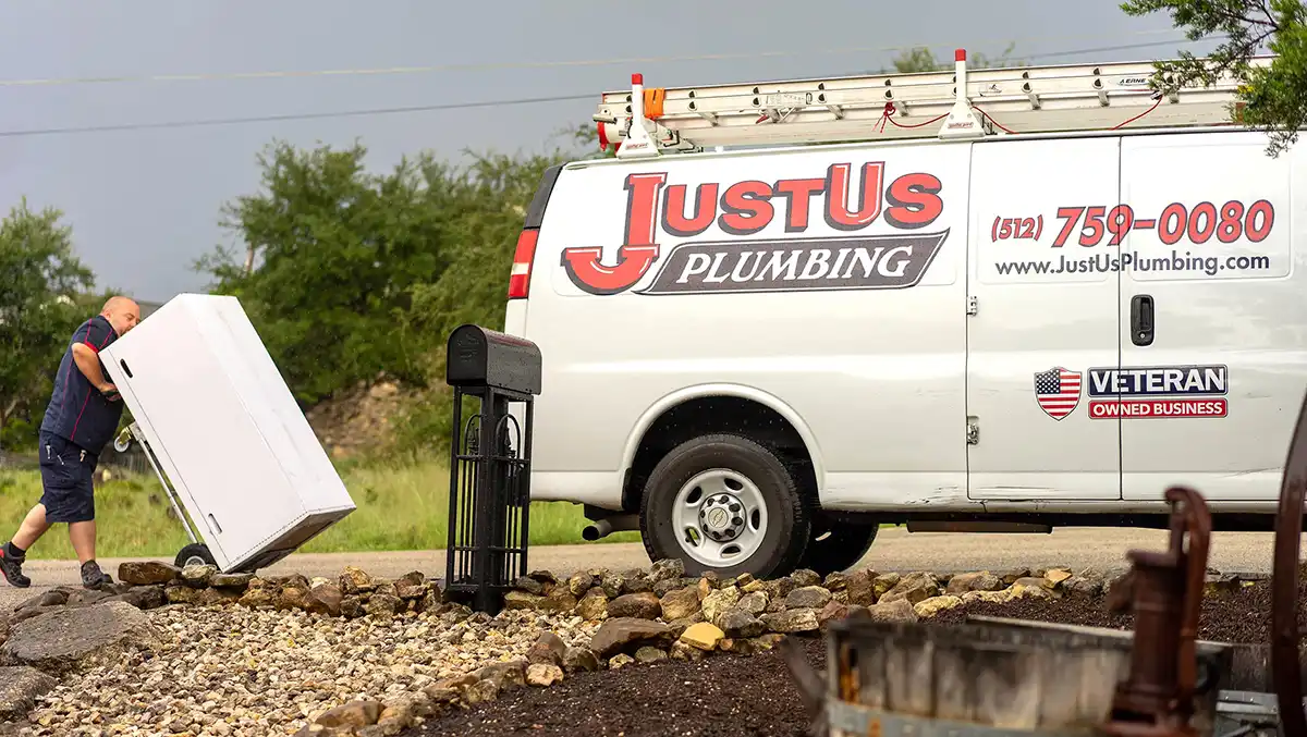 A man pushes a large white appliance on a dolly next to a white Just Us Plumbing service van parked on a road.
