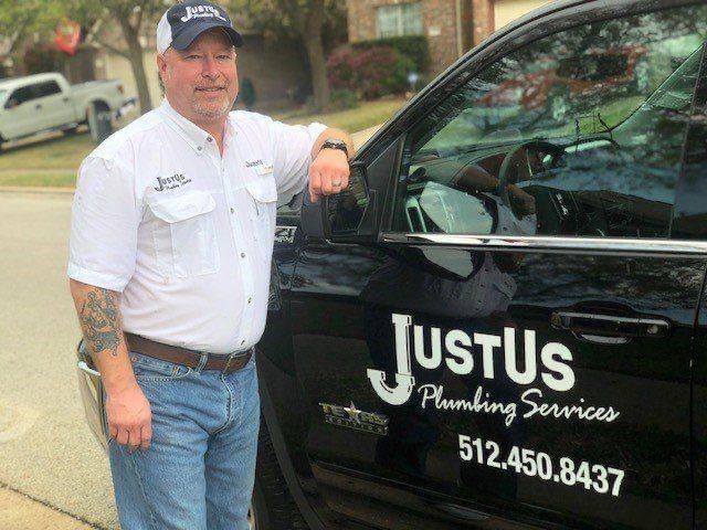 Man wearing a white JustUs Plumbing Services shirt and cap standing beside a black vehicle with the company logo and phone number 512.450.8437.